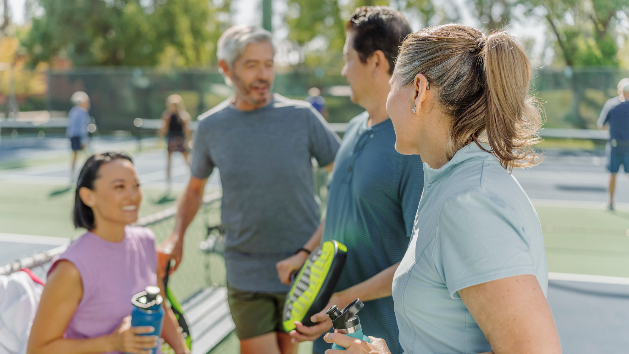 Menschen beim Tennis im Park, Halten Wasserflaschen, sprechen, im Hintergrund Tennisplätze, sonniges Wetter.