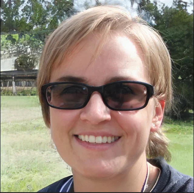 Close-up of a woman smiling outdoors, wearing dark sunglasses and a dark-colored top, with greenery and trees in the background.