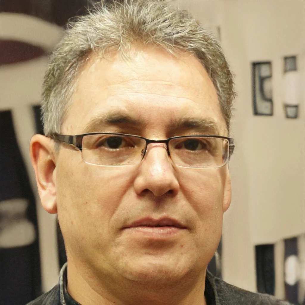 Close-up of a man with glasses and short gray hair, standing indoors with framed artwork in the background.