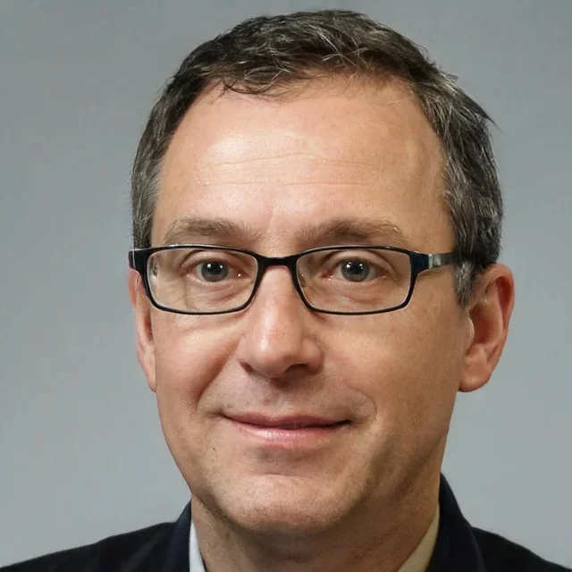 A man with short dark hair, glasses, and wearing a suit, smiling at the camera with a neutral gray background.