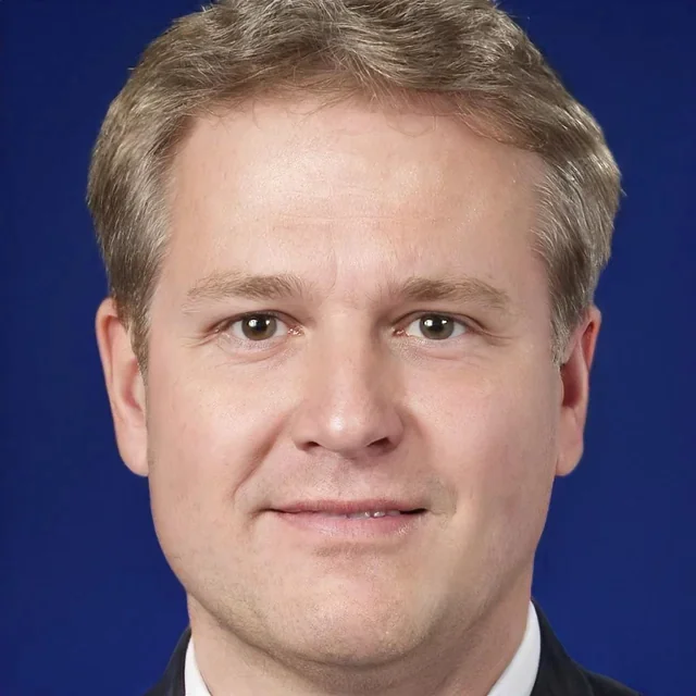 Headshot of a man with short blond hair, light skin, and a slight smile, wearing a suit and tie against a blue background.