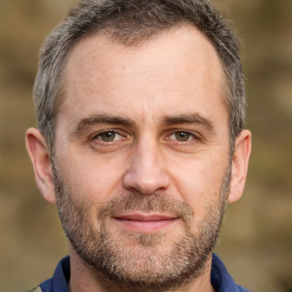 Close-up of a middle-aged man with short gray hair and a beard, wearing a dark blue shirt, outdoors with a blurred natural background.