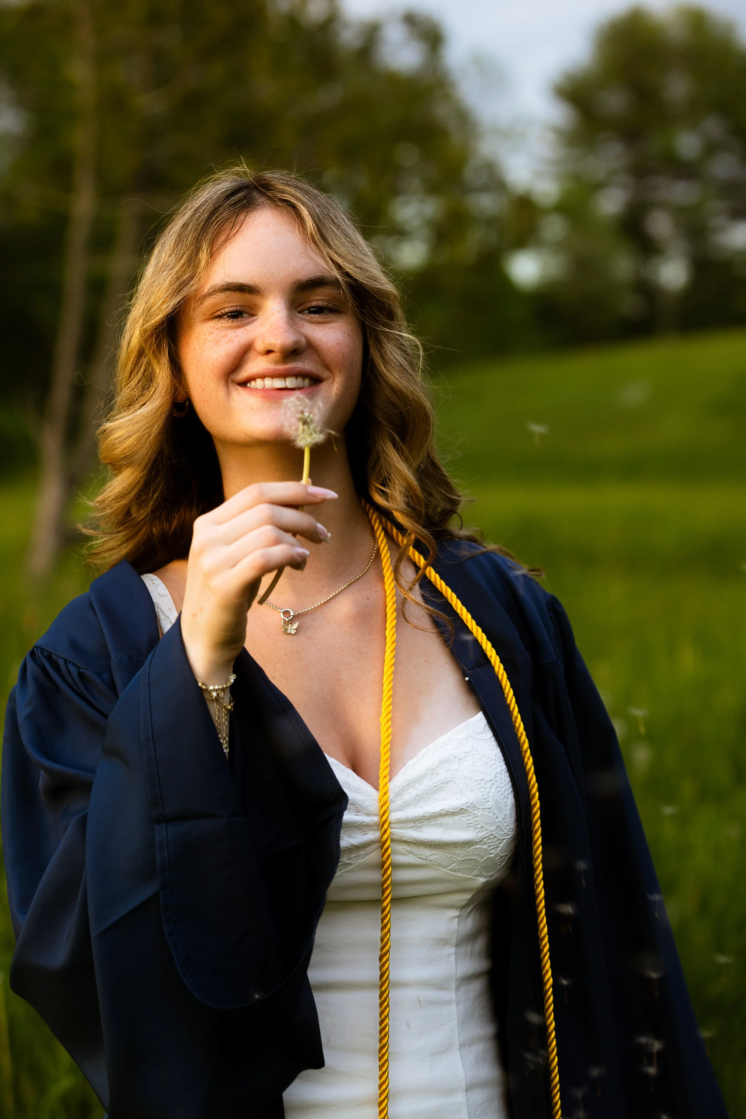 A young woman with wavy, shoulder-length brown hair, freckles, and hoop earrings, smiling outdoors during daytime, wearing a dark graduation gown with a yellow honor cord, holding a dandelion, with blurred green trees in the background.