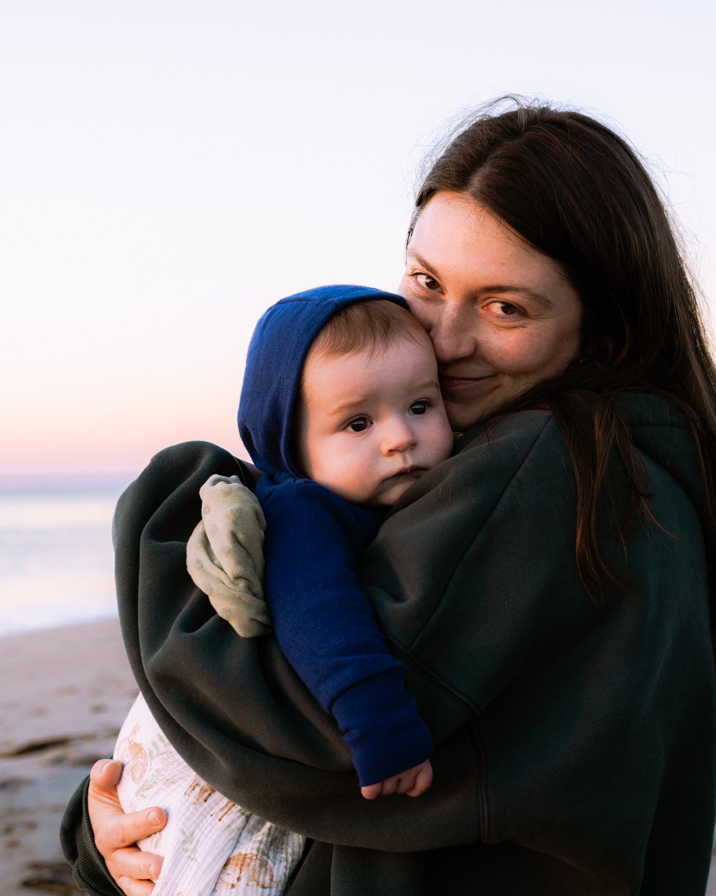 A woman with long brown hair and a black jacket hugs and kisses two children on a beach at sunset, with a calm ocean and sky in the background.