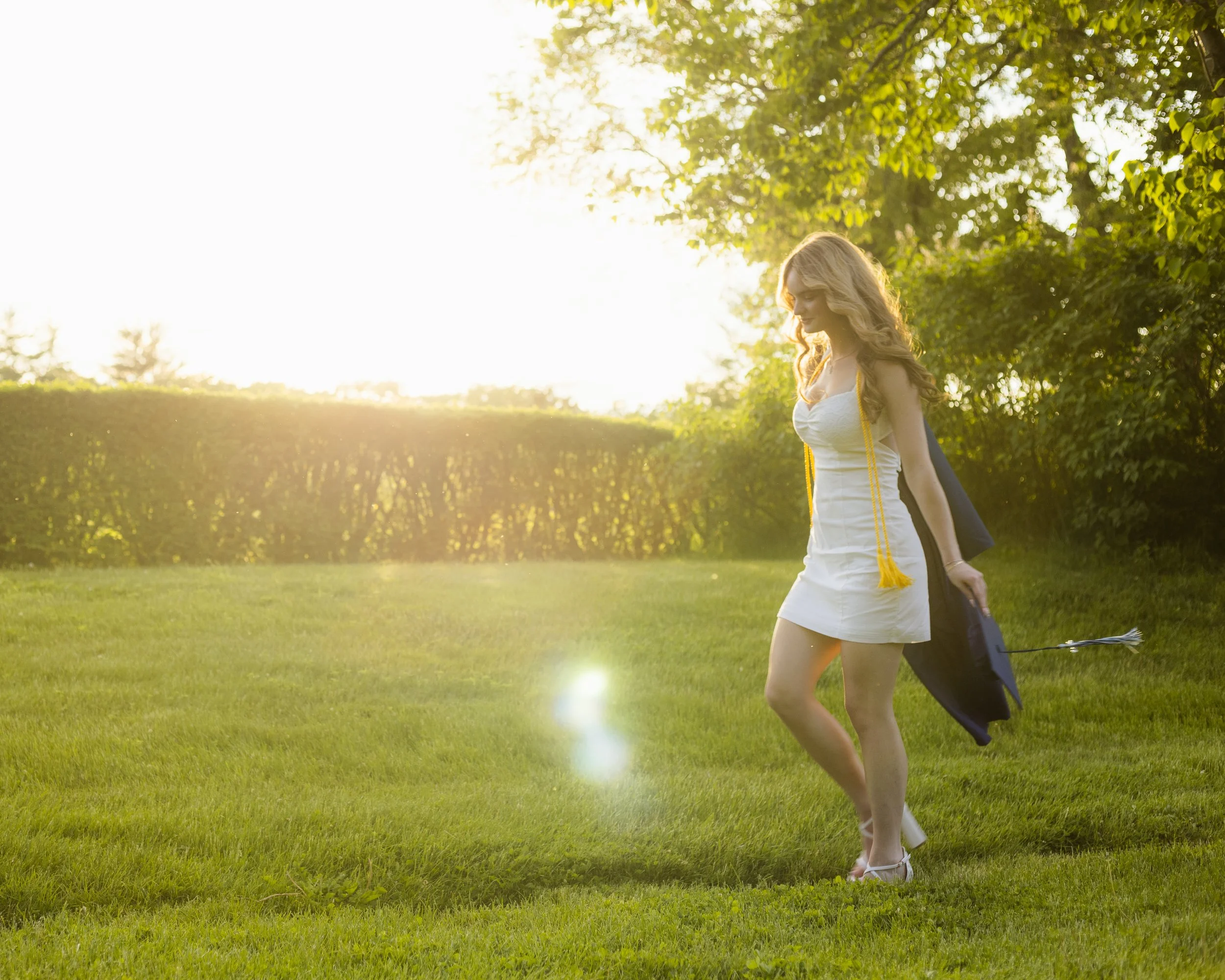 A young woman in a white dress walking on grass in a park during sunset, carrying a black golf bag with clubs.