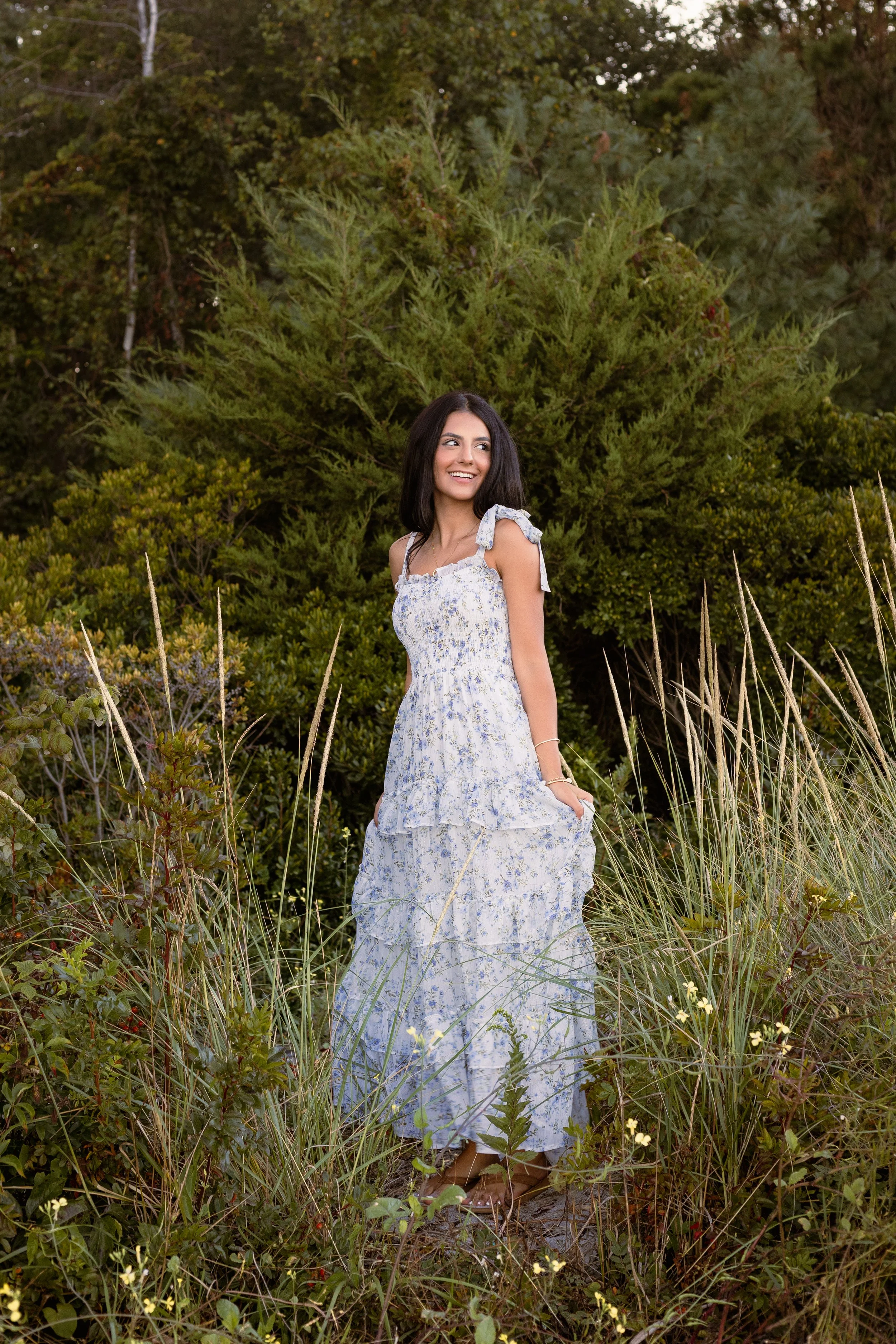 A young woman with dark hair wearing a white floral dress standing in a lush field of tall grasses and wildflowers, with dense green bushes and trees in the background, smiling and looking to the side.