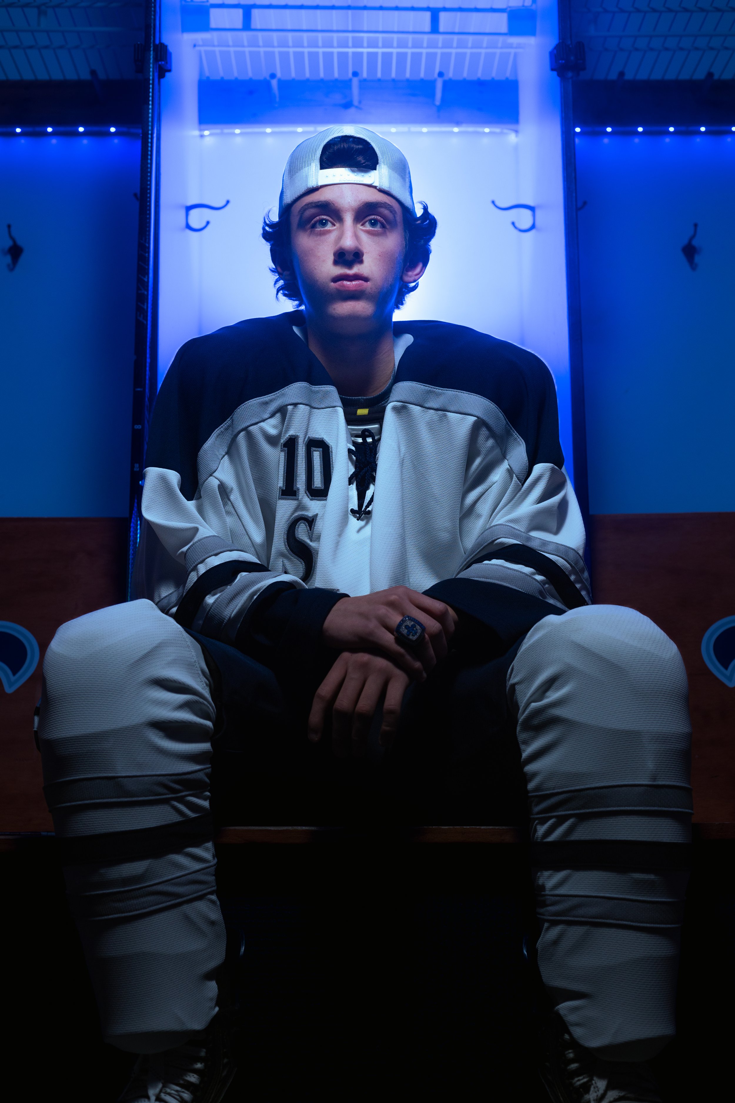 Young male hockey player sitting on bench in locker room, wearing jersey, hockey gloves, and cap, illuminated by blue lighting.