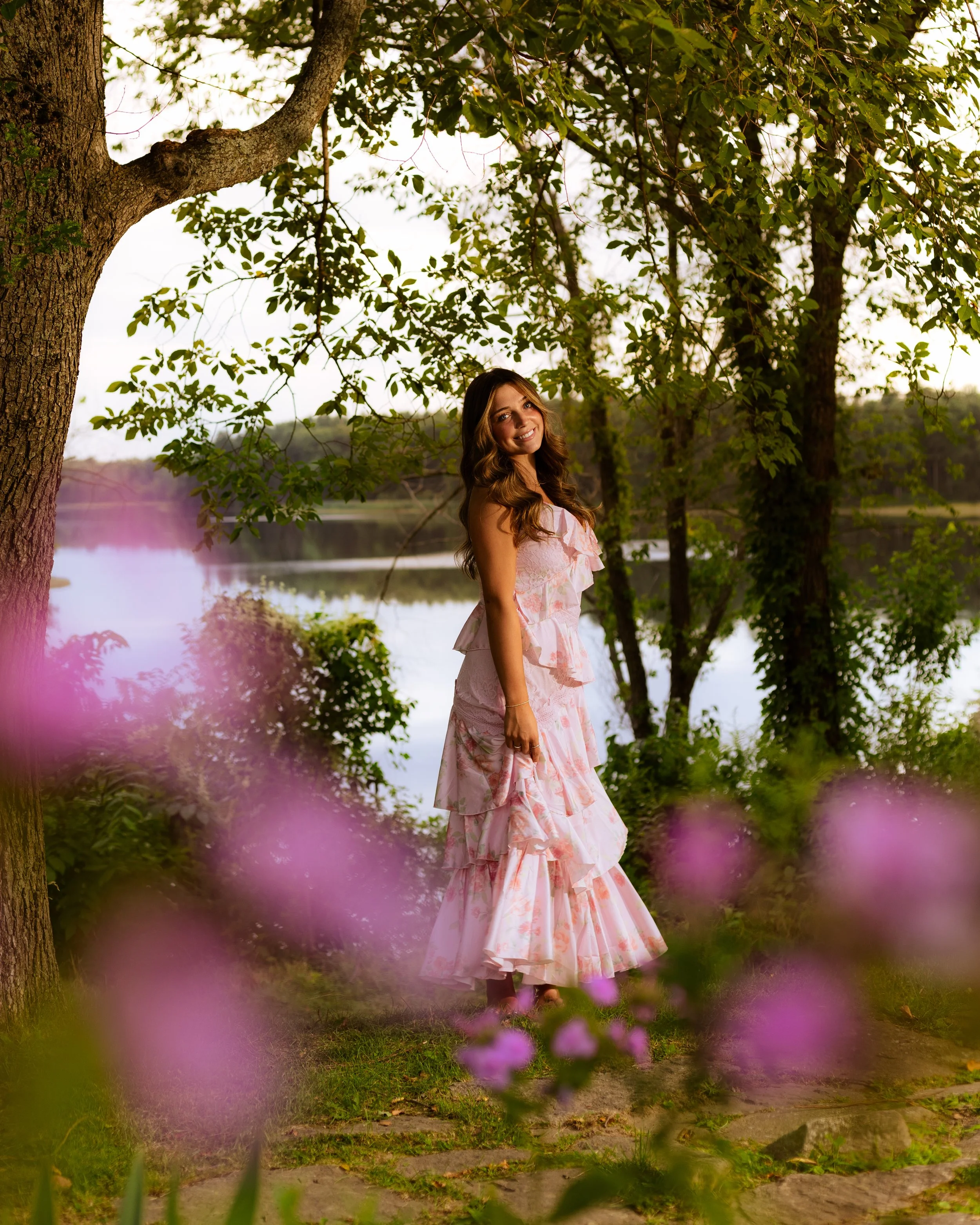 A woman in a pink floral dress smiling outdoors near a large tree and a body of water, with pink flowers in the foreground.