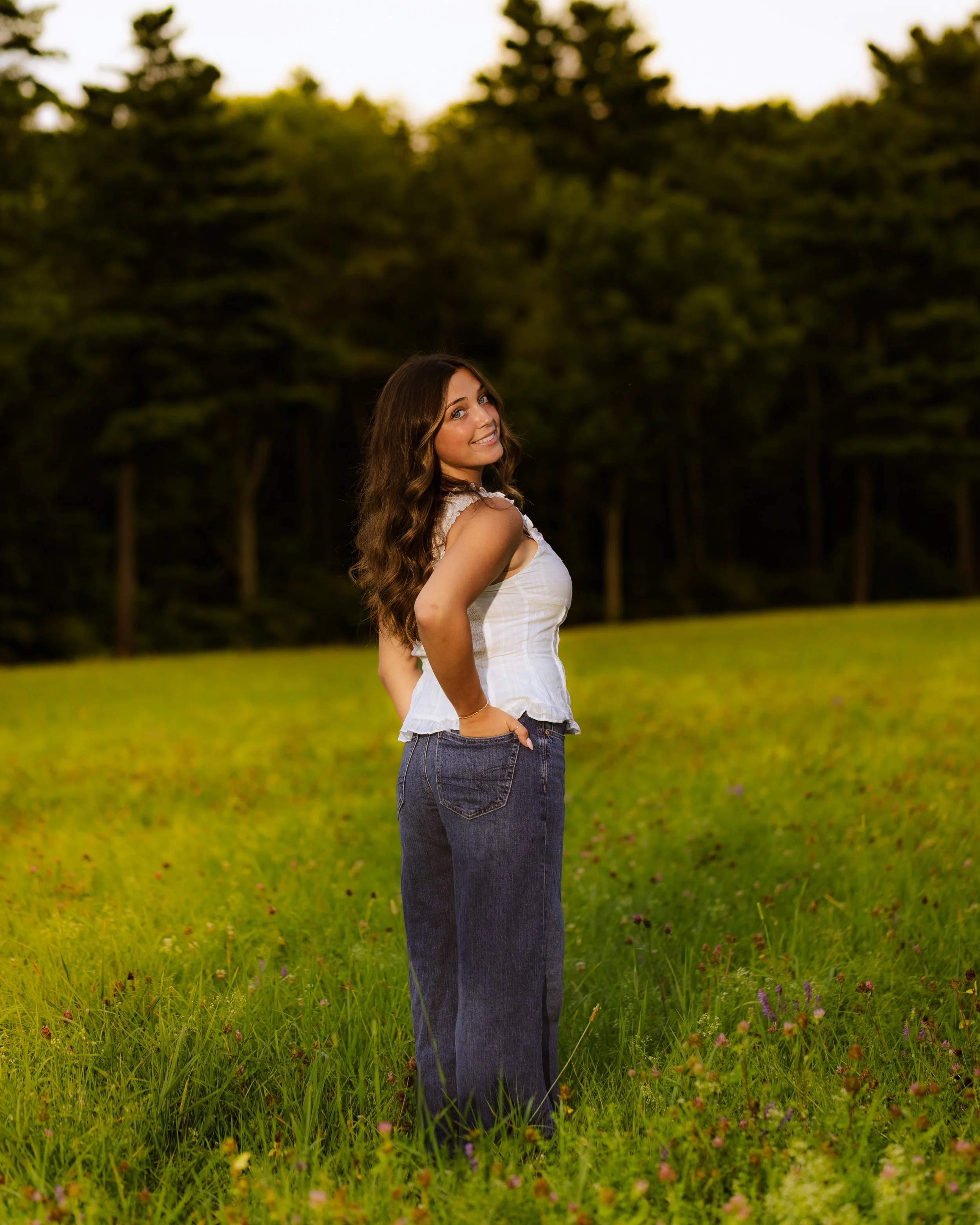 A woman with long brown hair smiling and looking back over her shoulder in a white sleeveless top and blue jeans, standing in a lush green field with a line of trees in the background.