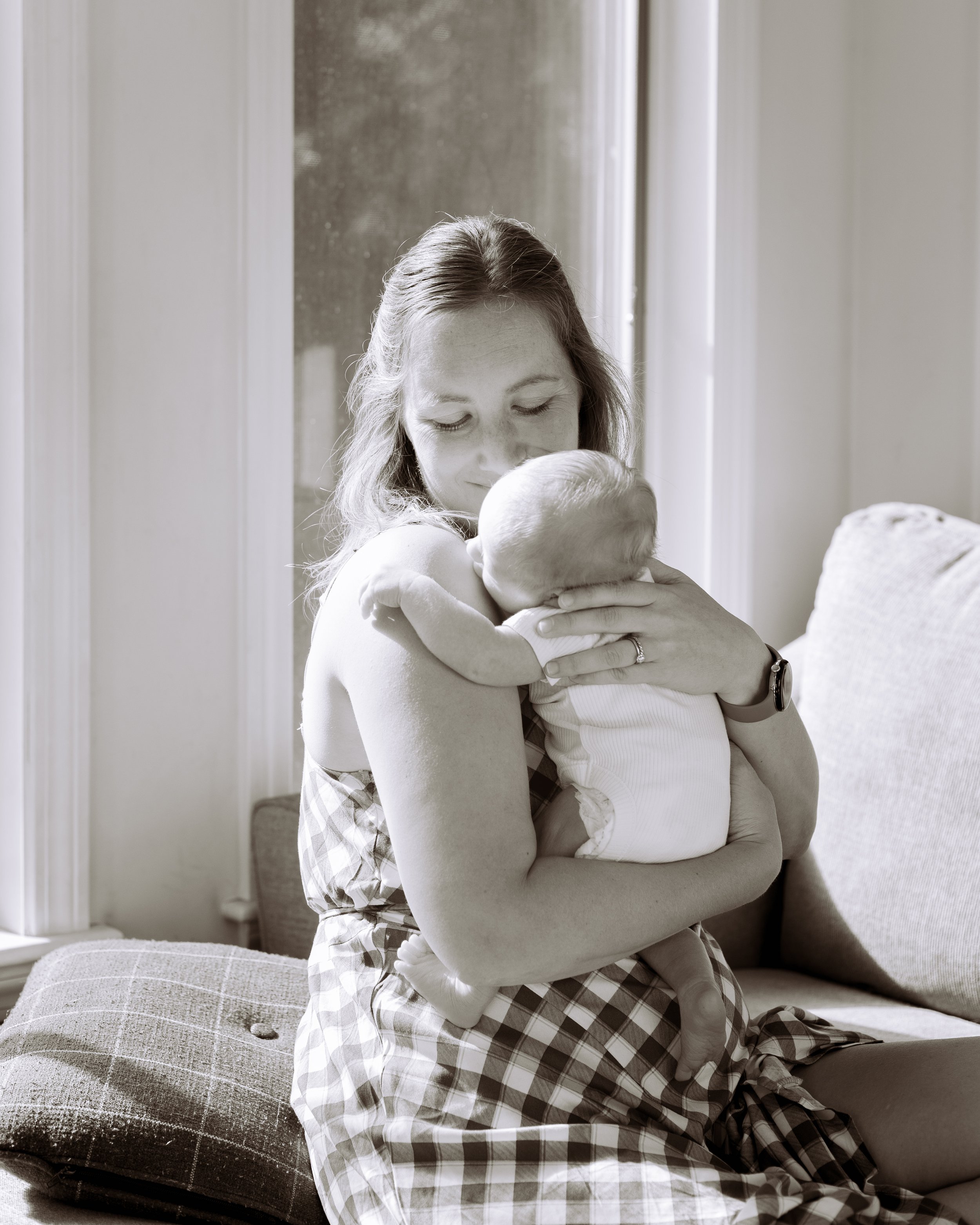 A woman holding and embracing a small baby, both indoors near a window, sitting on a cushioned surface.
