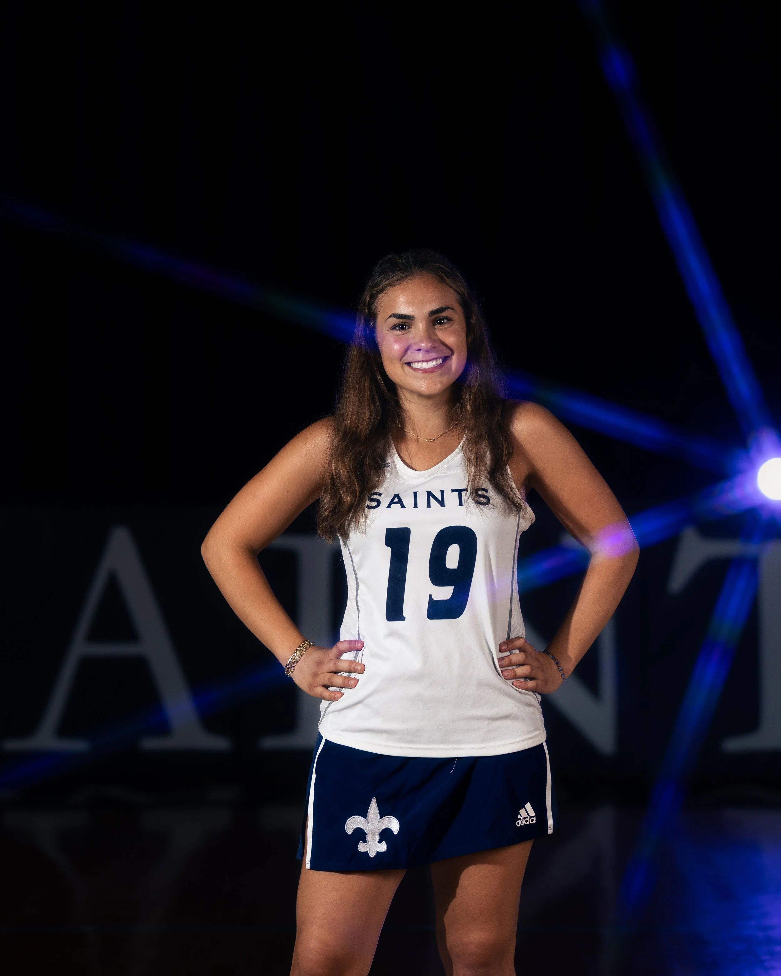 A young woman smiling confidently with hands on hips, wearing a white sports jersey with 