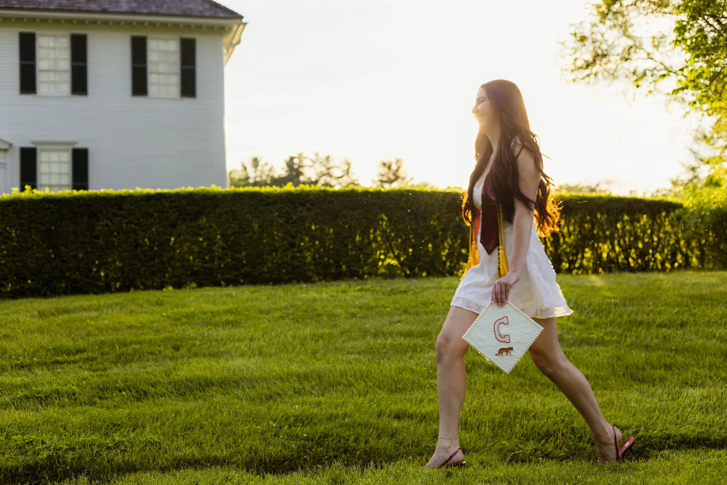 A young woman with long hair wearing a white dress and sandals is walking on grass during sunset. She is holding a decorated diploma or certificate with the letter 'C' and a bear illustration. There are trees and a white building in the background.