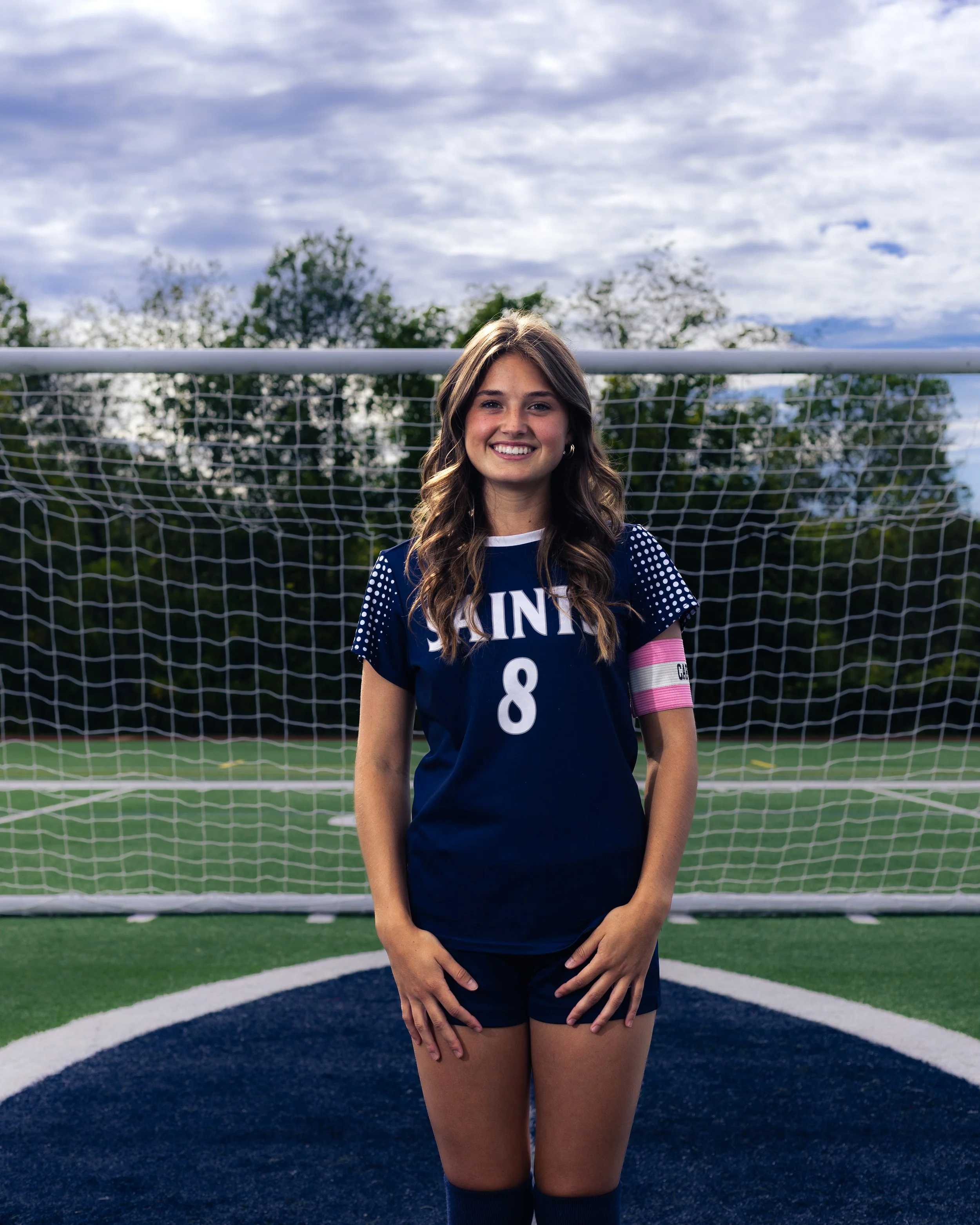 Young woman wearing a navy sports uniform with the number 8, kneeling in front of a soccer goal on a green field, with trees and cloudy sky in the background.