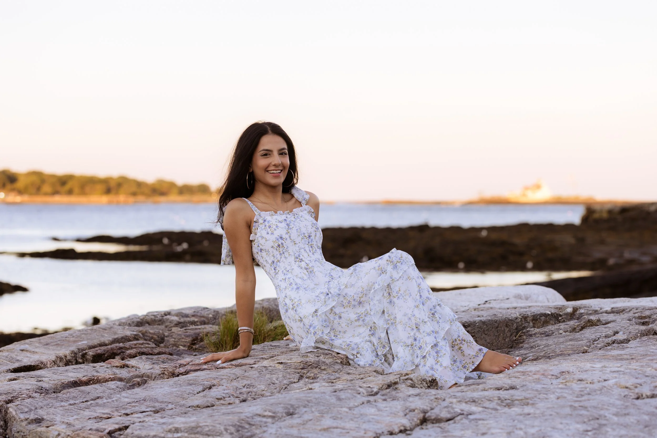 A woman in a white floral dress sitting on rocks near the water at sunset, smiling at the camera.