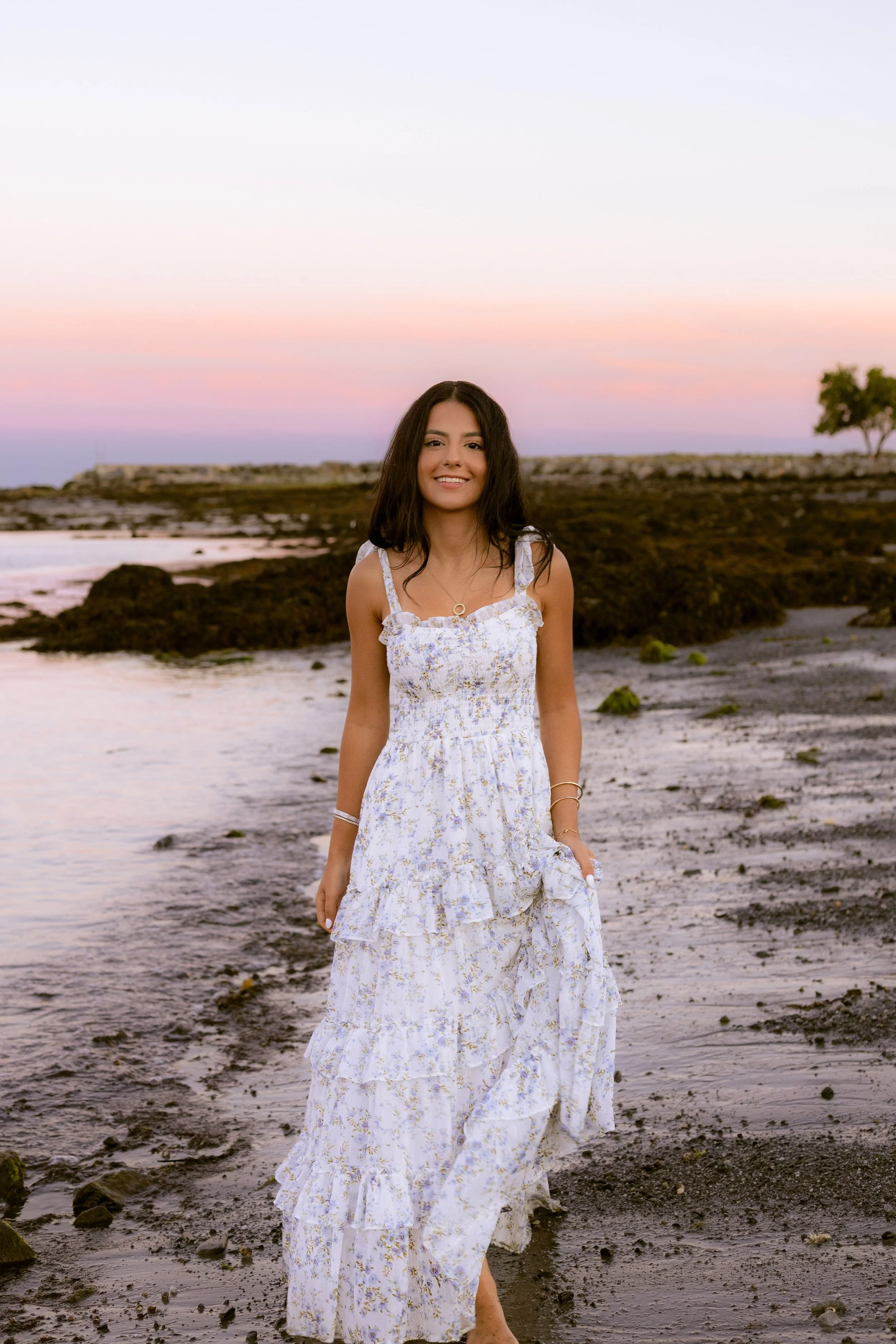 A woman in a white floral maxi dress standing on a beach at sunset with a colorful sky in the background, smiling and holding her dress.