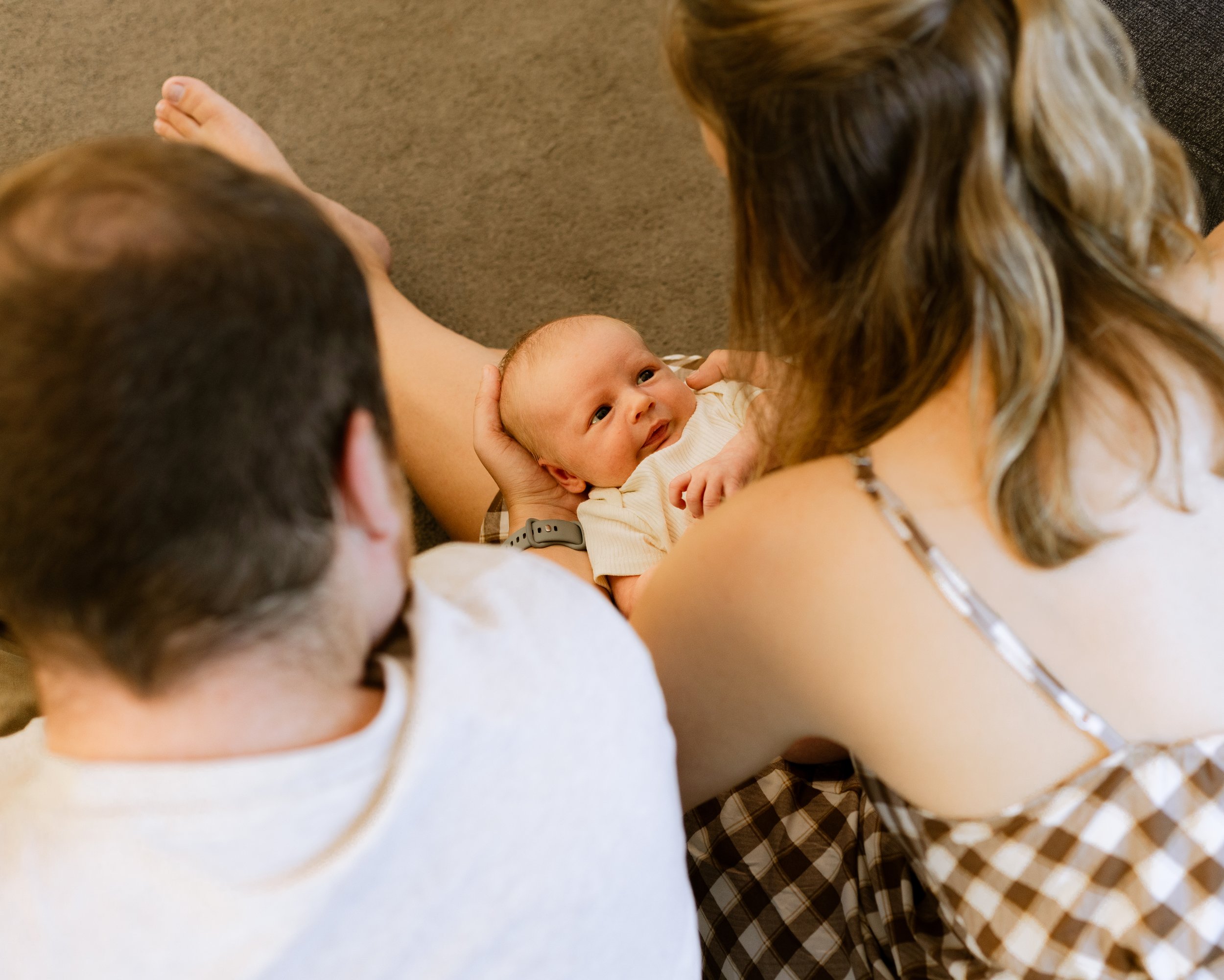 A woman lying on the floor looking up at a baby in her arms, surrounded by two men, all appearing to be in a relaxed setting.