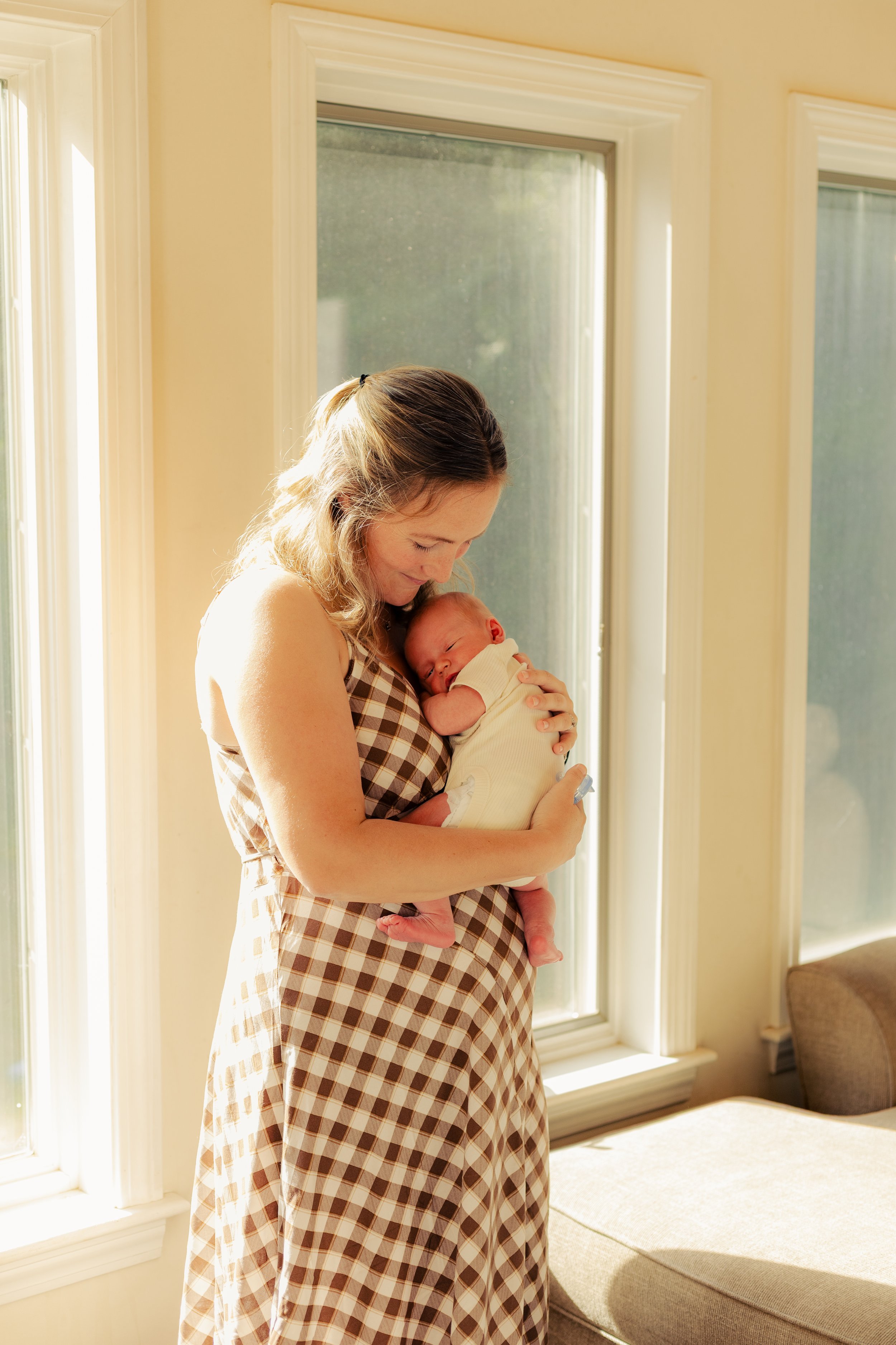 A woman holds a newborn baby near a window in a sunlit room, smiling softly.