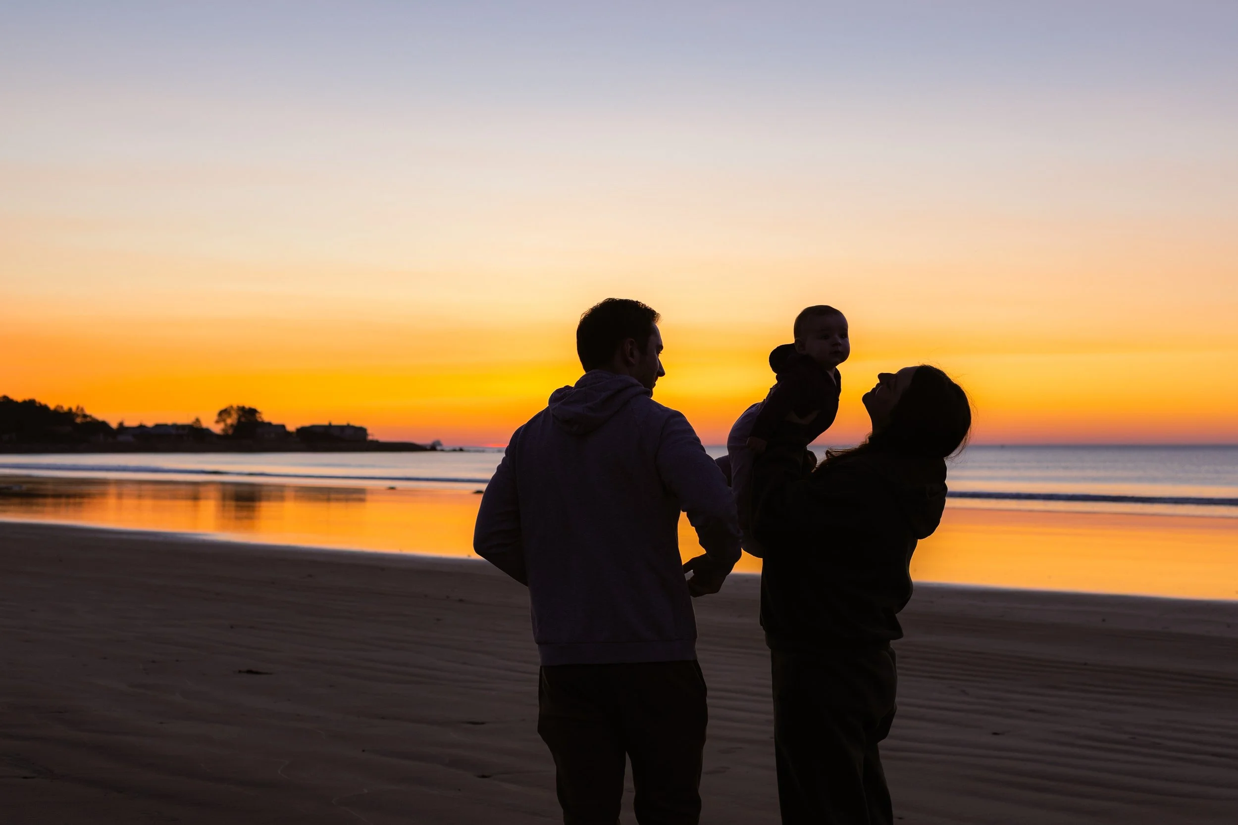 A family of three at the beach during sunset, with the parents holding their child between them.
