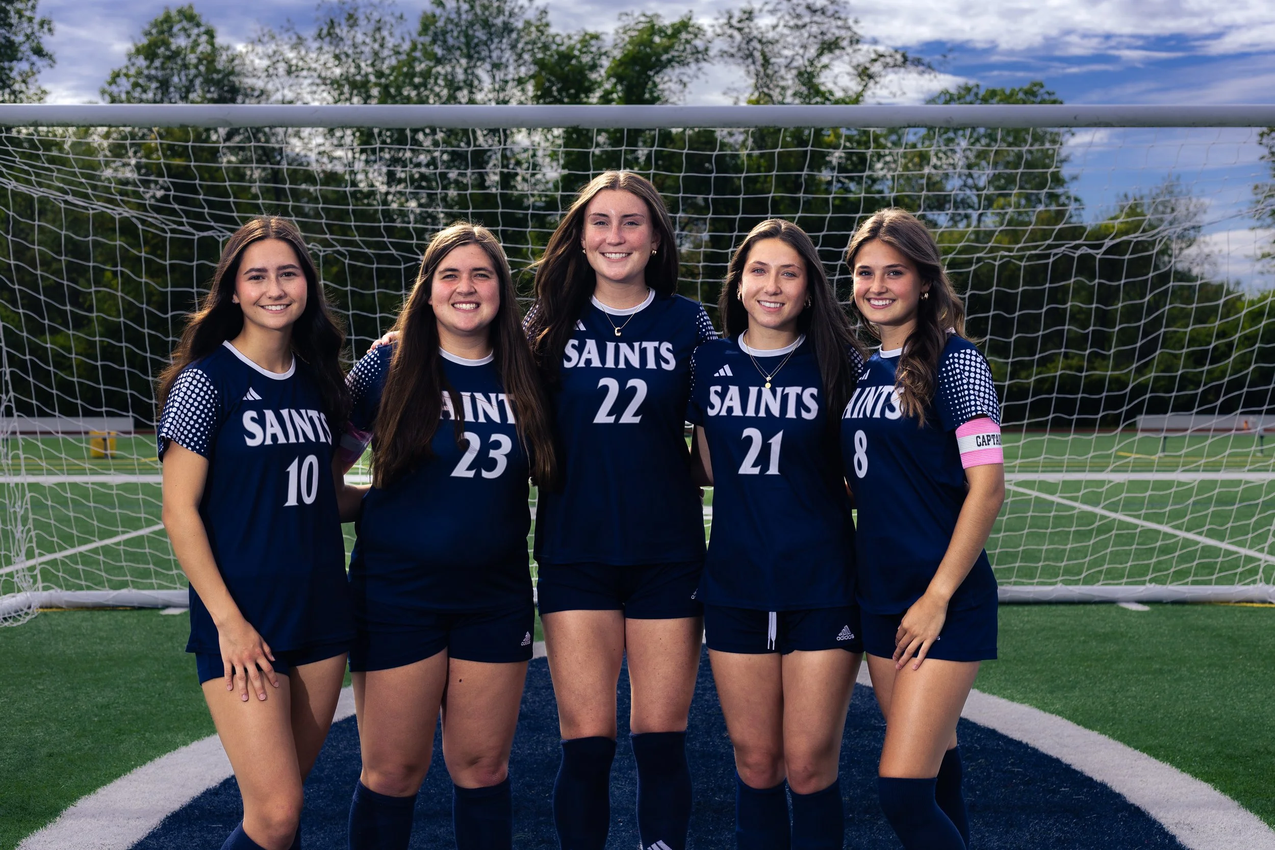 A group of five young women in navy blue soccer uniforms standing together on a soccer field in front of a goal net, smiling for the camera.