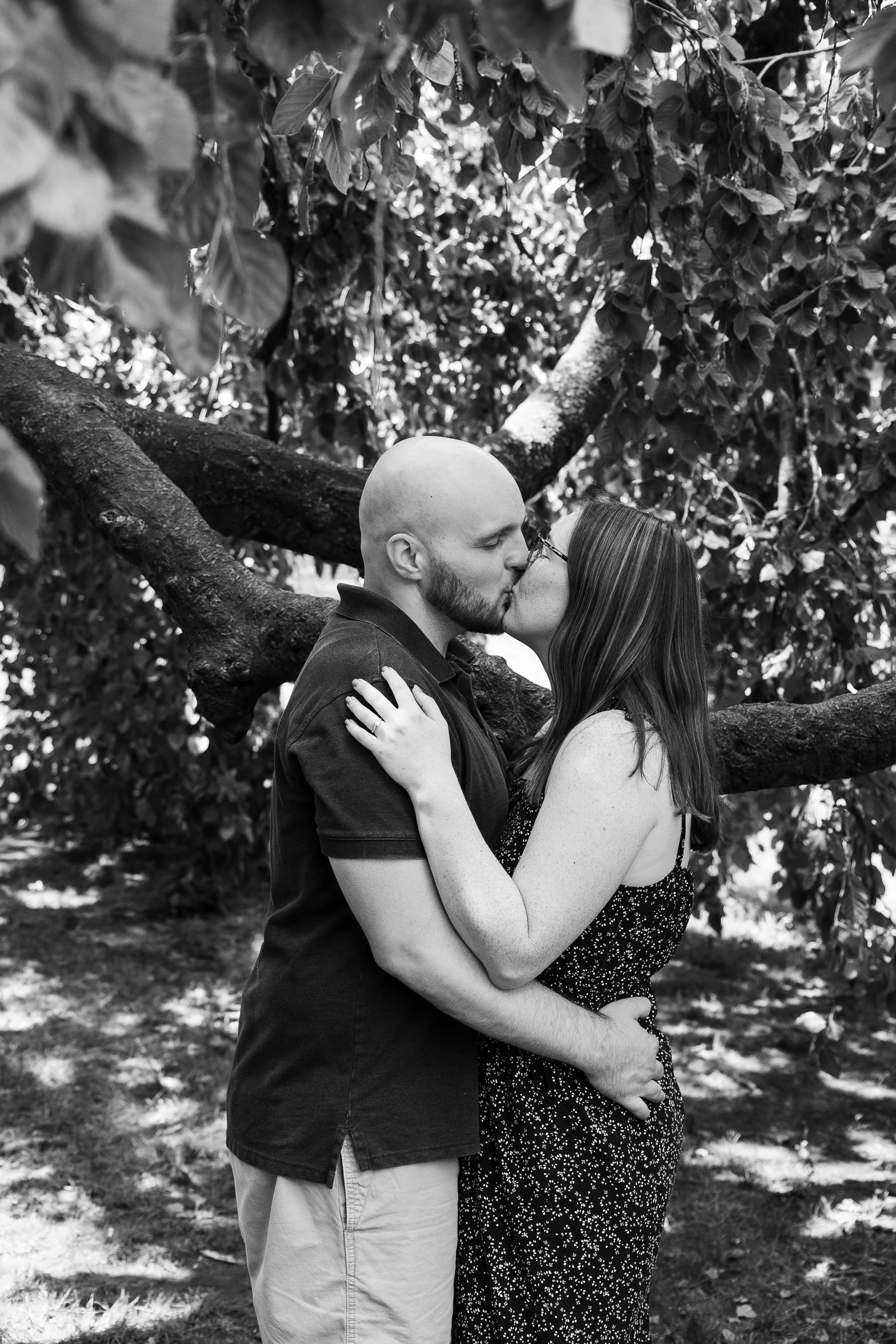 A couple kissing outdoors in front of a large tree with thick branches, black and white photograph.
