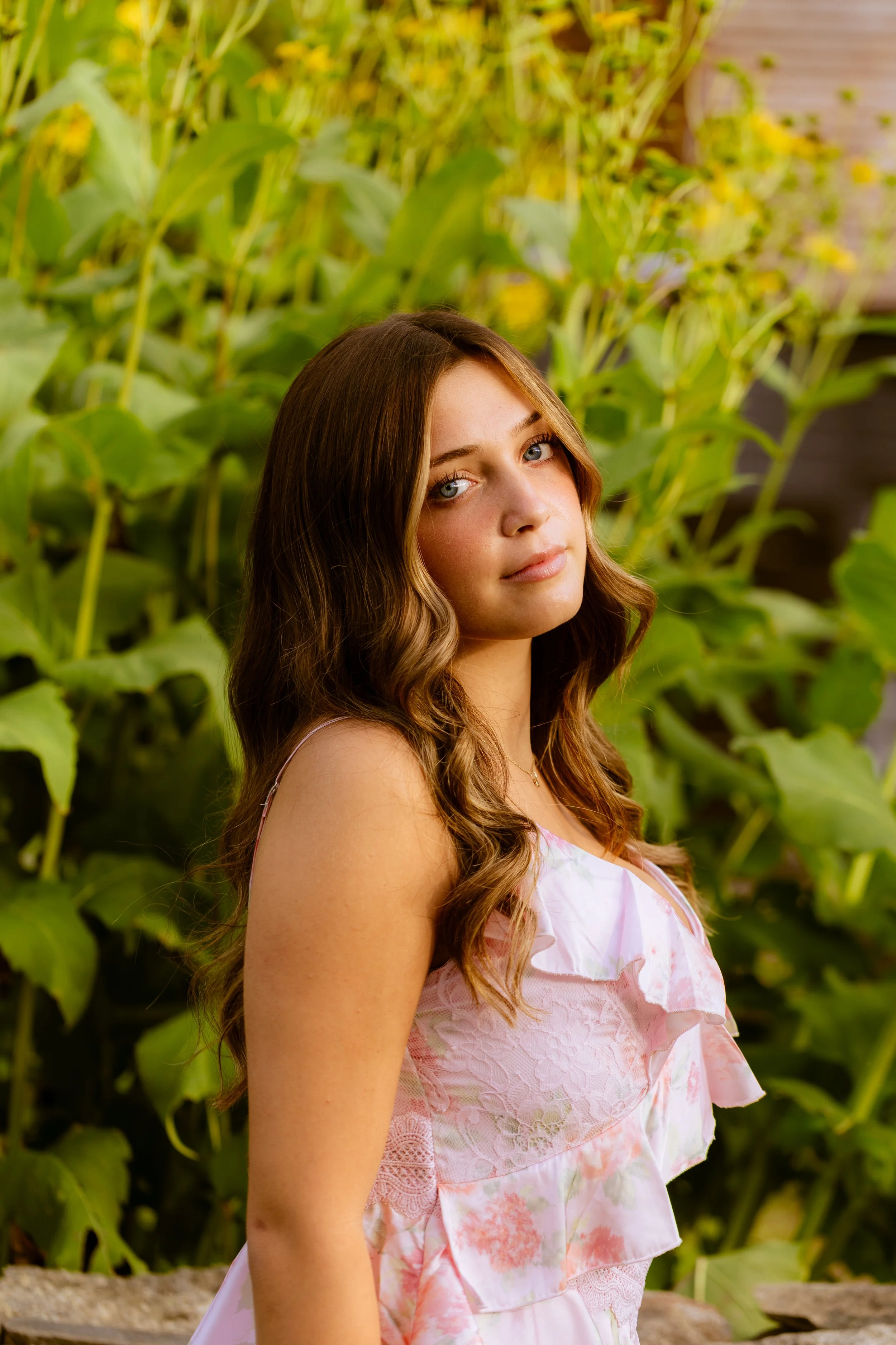 A young woman with long wavy brown hair and blue eyes wearing a pink and white floral dress standing outdoors among green plants.