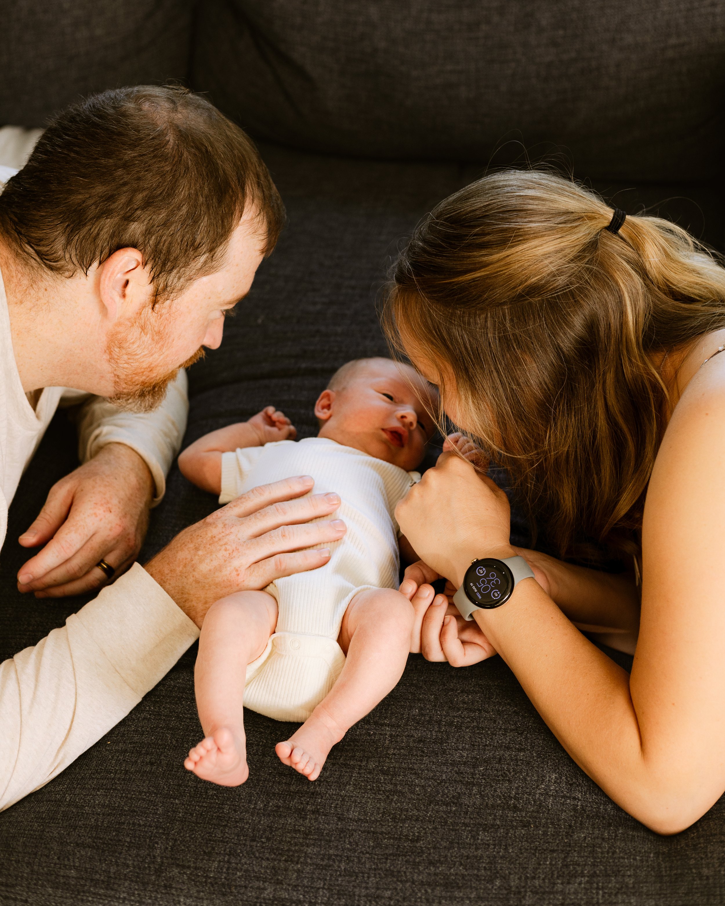 A couple with a baby lying on a black couch, leaning over and looking at the baby, with the man on the left and the woman on the right. The woman is wearing a smartwatch, and the baby is wearing a white outfit.