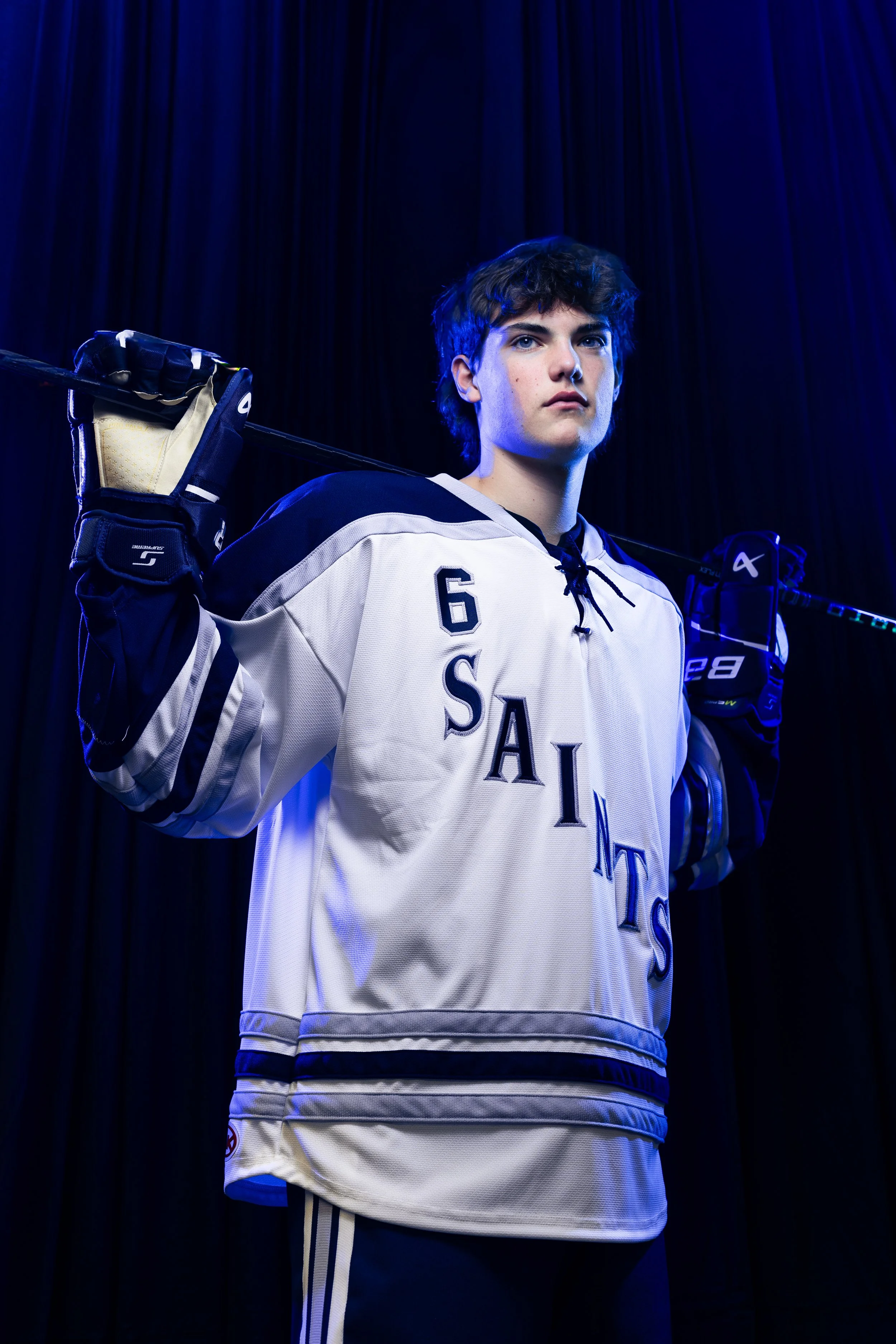 Young male ice hockey player standing with hockey stick over shoulders, wearing white jersey with 'SAINTS' and the number 6, black gloves, and dark pants, against dark curtain background.