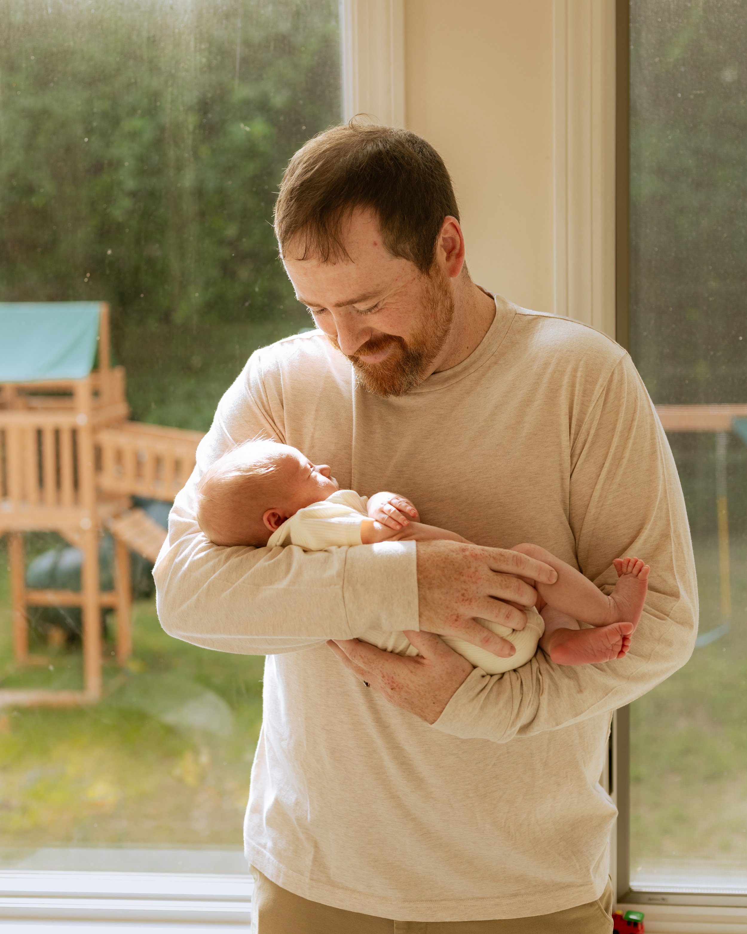 A man with a beard and light brown hair holds a newborn baby by a window inside a house. The man looks down at the baby, who is wrapped in a blanket. There is a window with a view of a green yard outside.