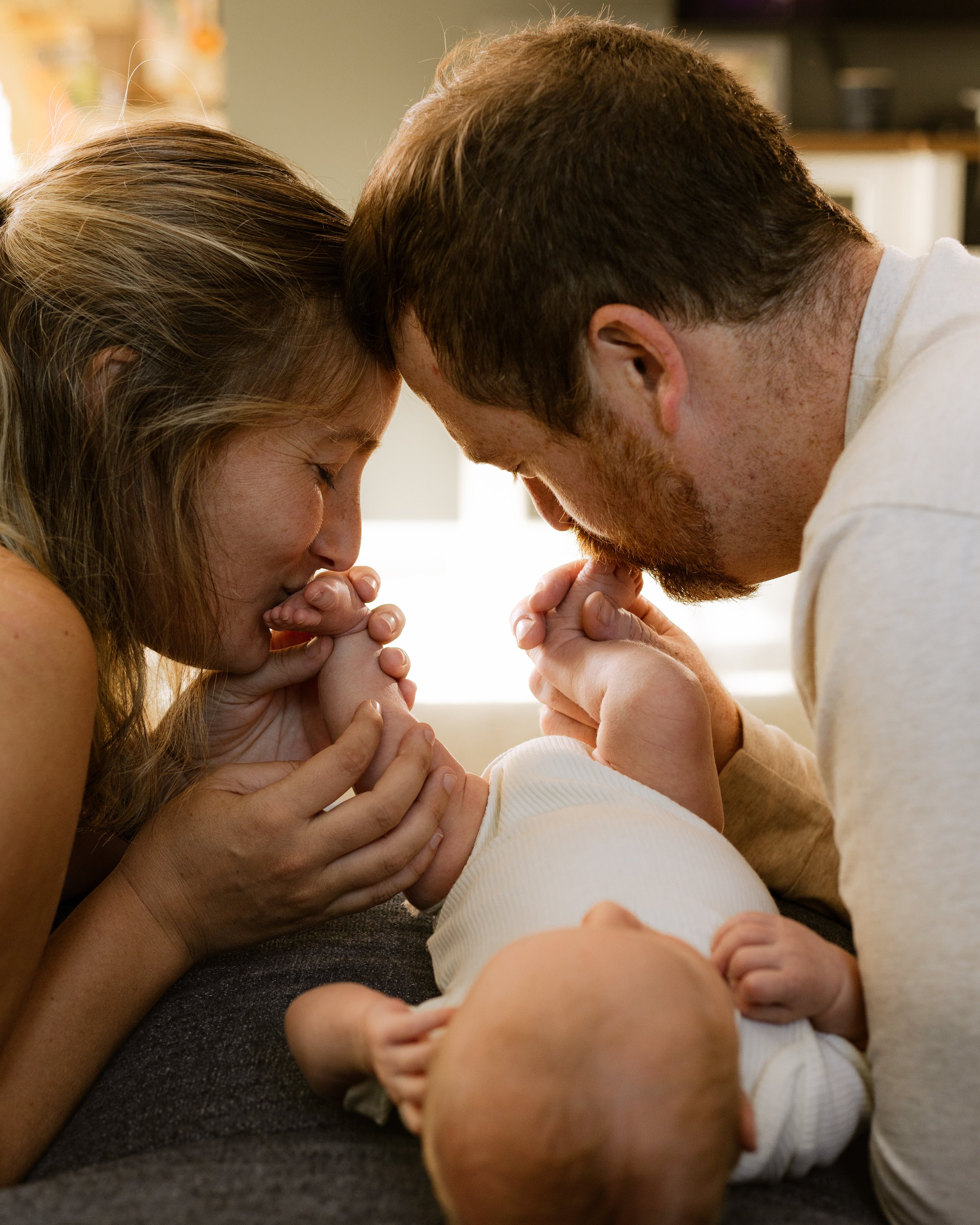A family, including a man, woman, and baby, are engaging in a close, intimate moment, with the adults and baby touching foreheads and holding hands in a warm, affectionate setting.
