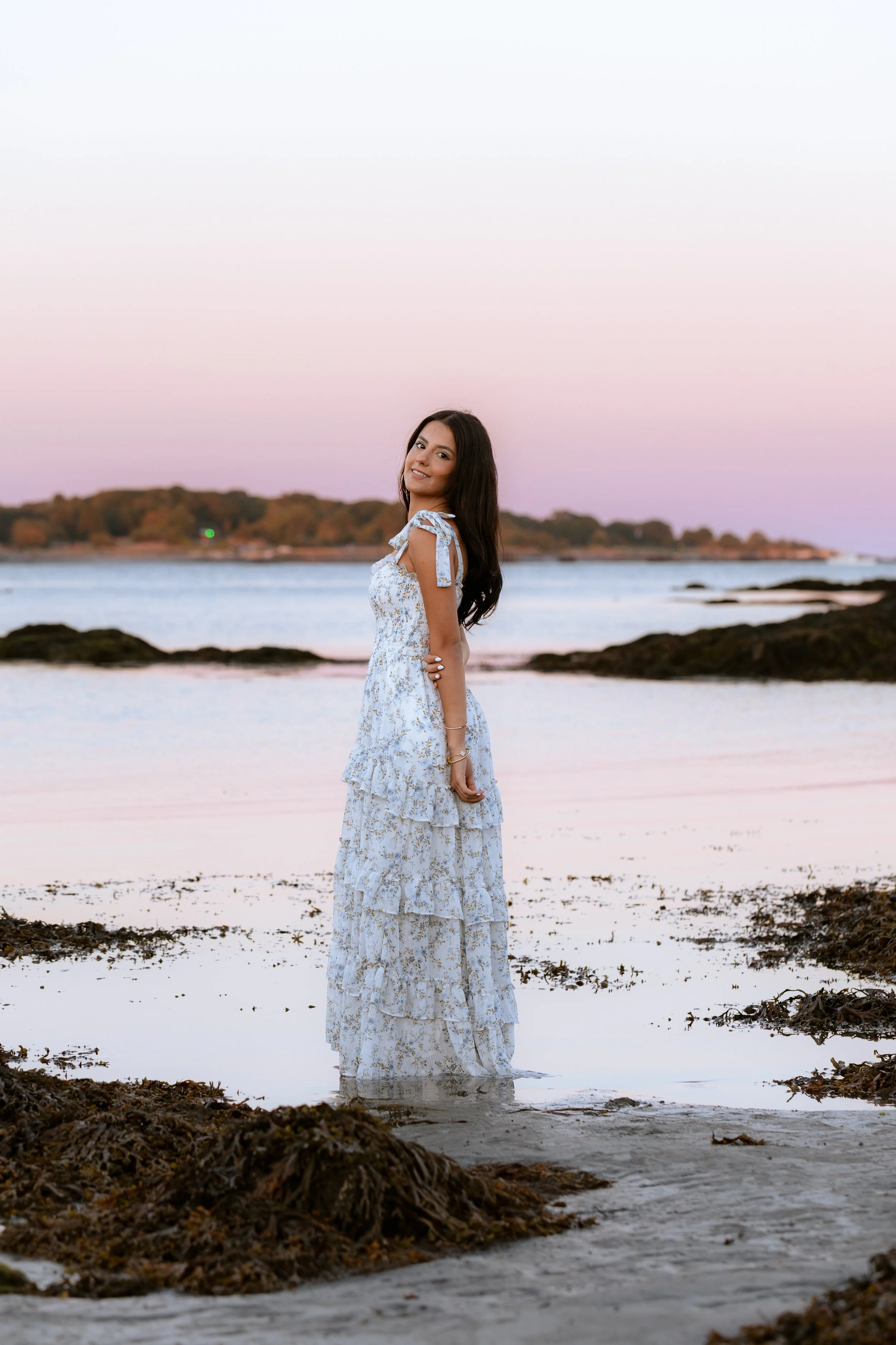 A woman in a flowy, floral white sundress is standing in shallow water at the beach during sunset, with the sky displaying pink and purple hues and a distant shoreline with trees in the background.
