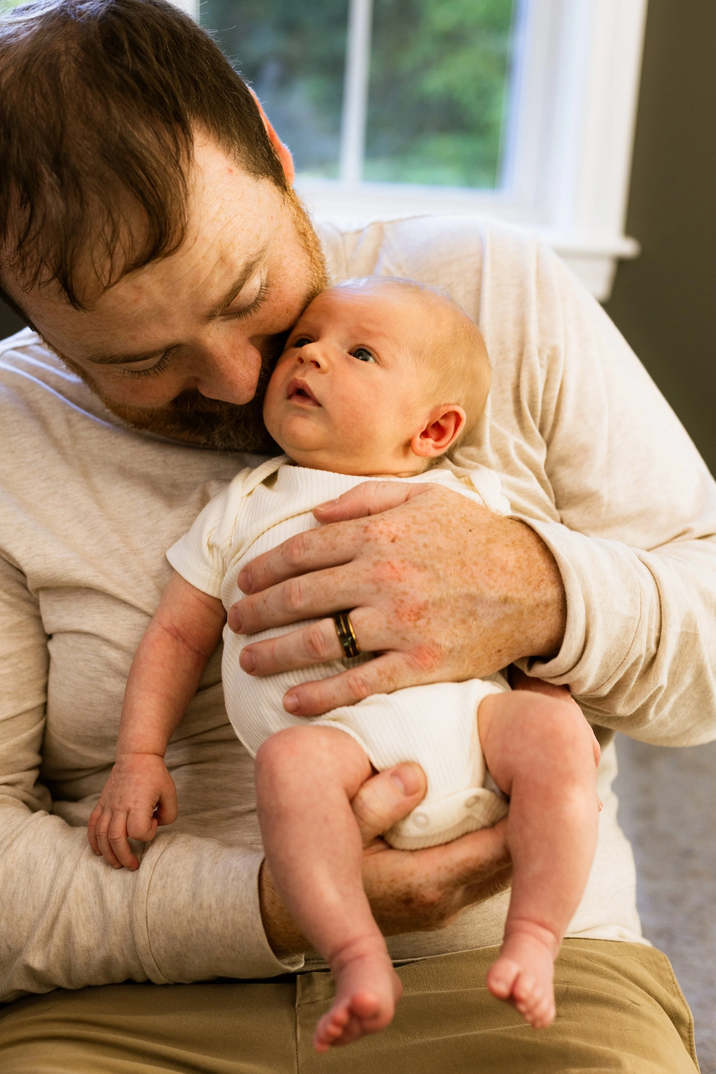 A man with a beard and short brown hair holding a baby with light skin, blond hair, and blue eyes, near a window with greenery outside.