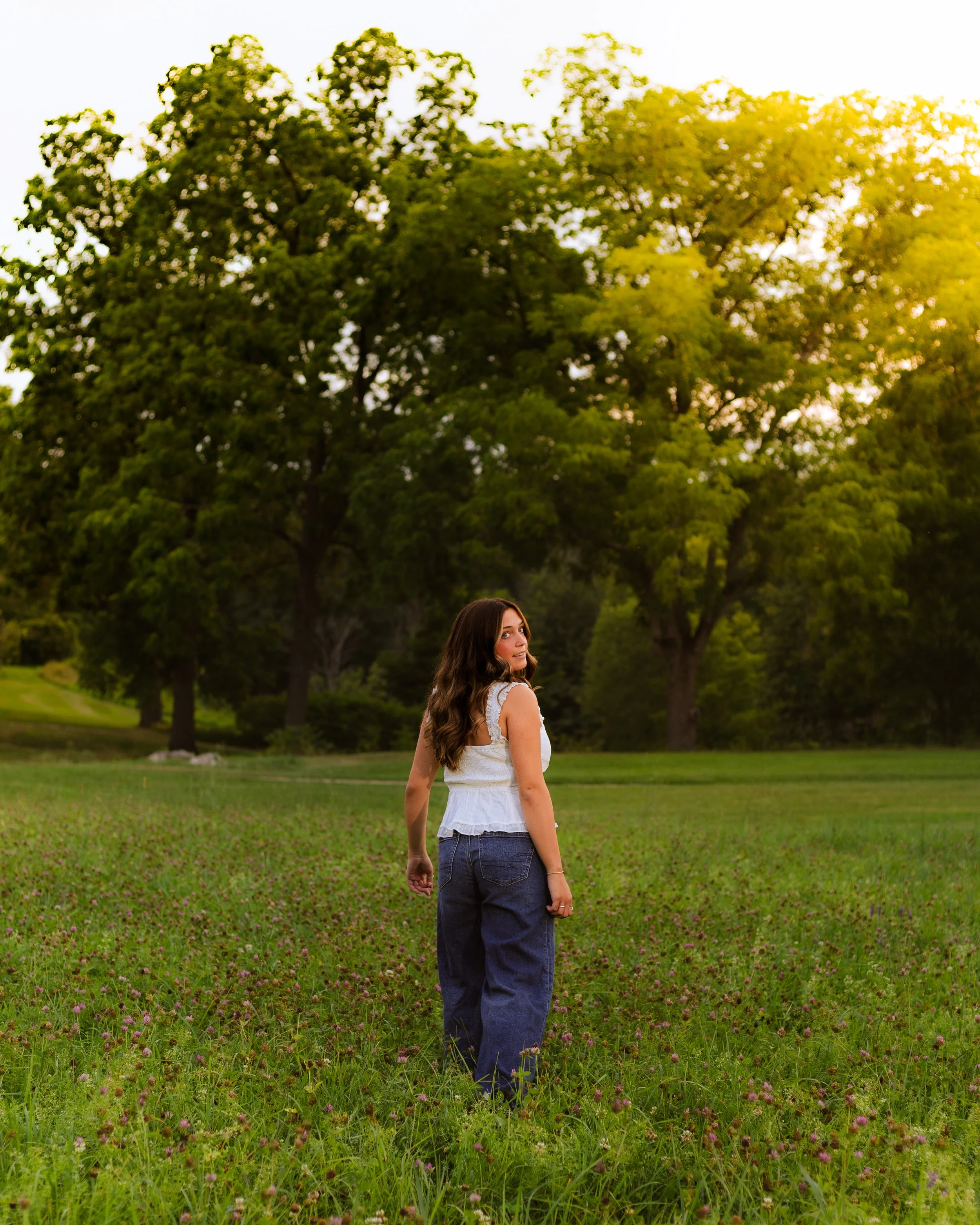 A young woman with long brown hair, wearing a white sleeveless top and dark pants, standing in a field of green grass and small purple flowers, with tall trees and a bright sky in the background.