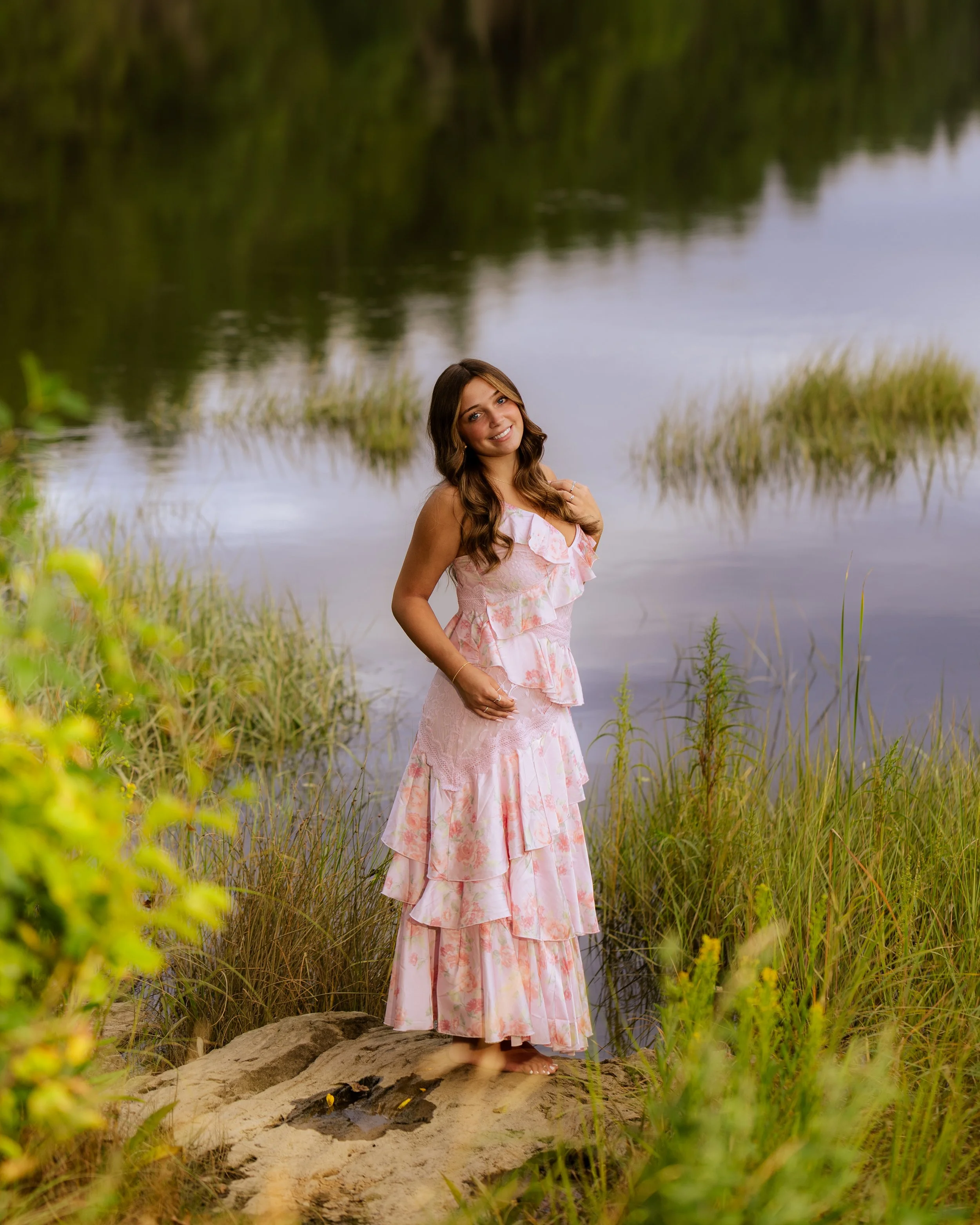 A woman standing on a rock at the edge of a body of water, surrounded by grass and greenery, smiling at the camera.