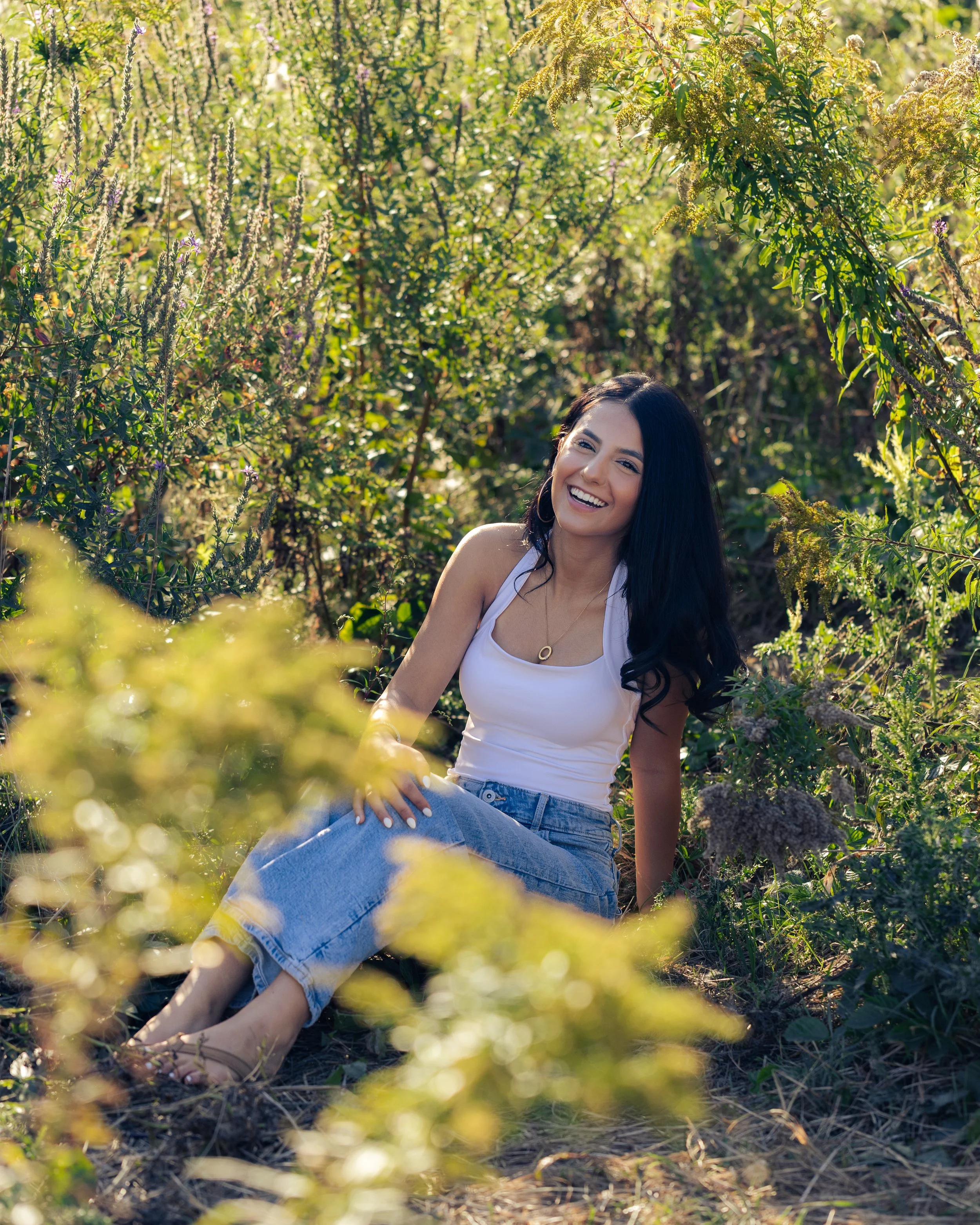 A young woman with long dark hair sitting outdoors in a lush garden of green plants, smiling and enjoying the sunshine.