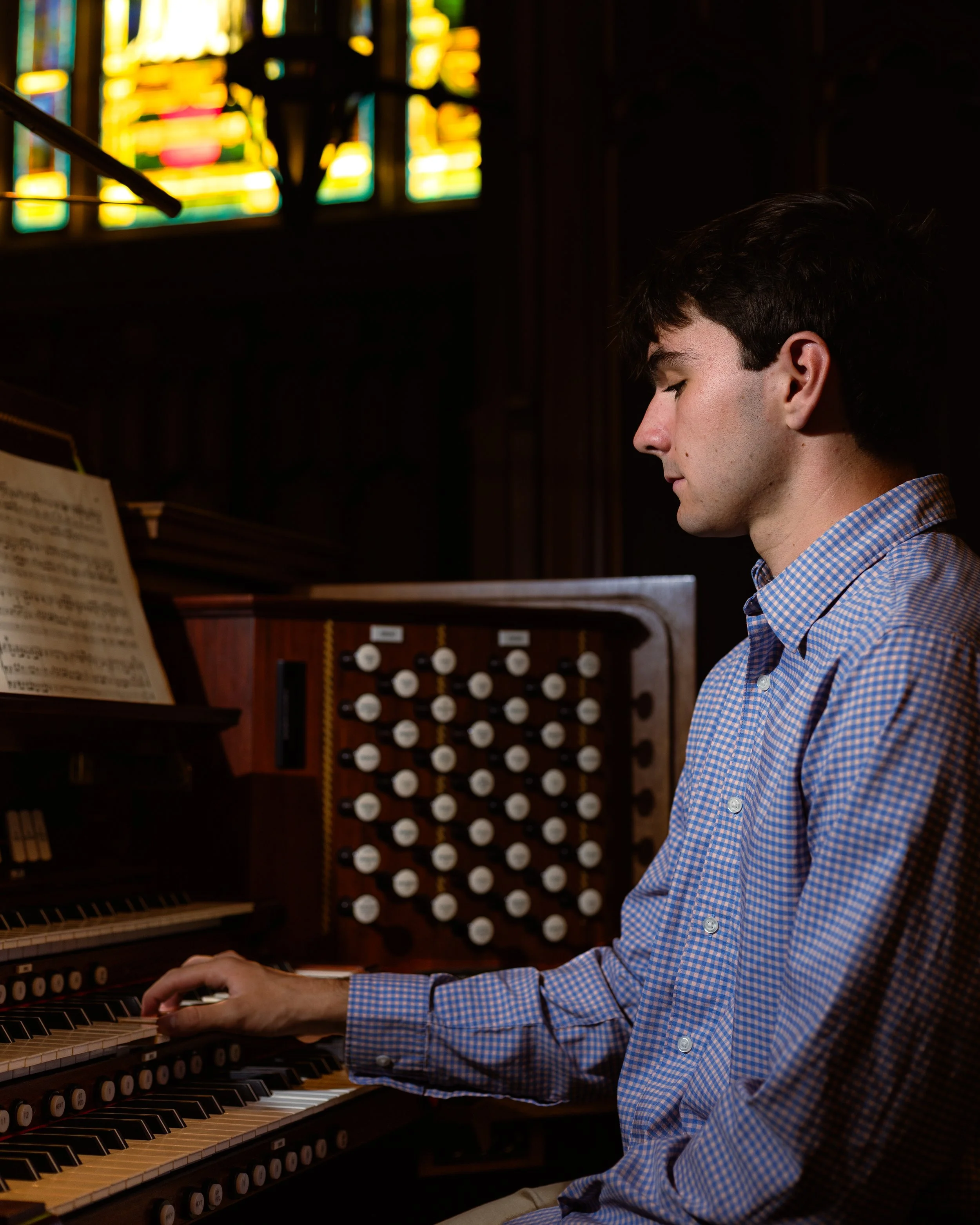 Young man playing a pipe organ inside a church or cathedral with stained glass windows in the background.