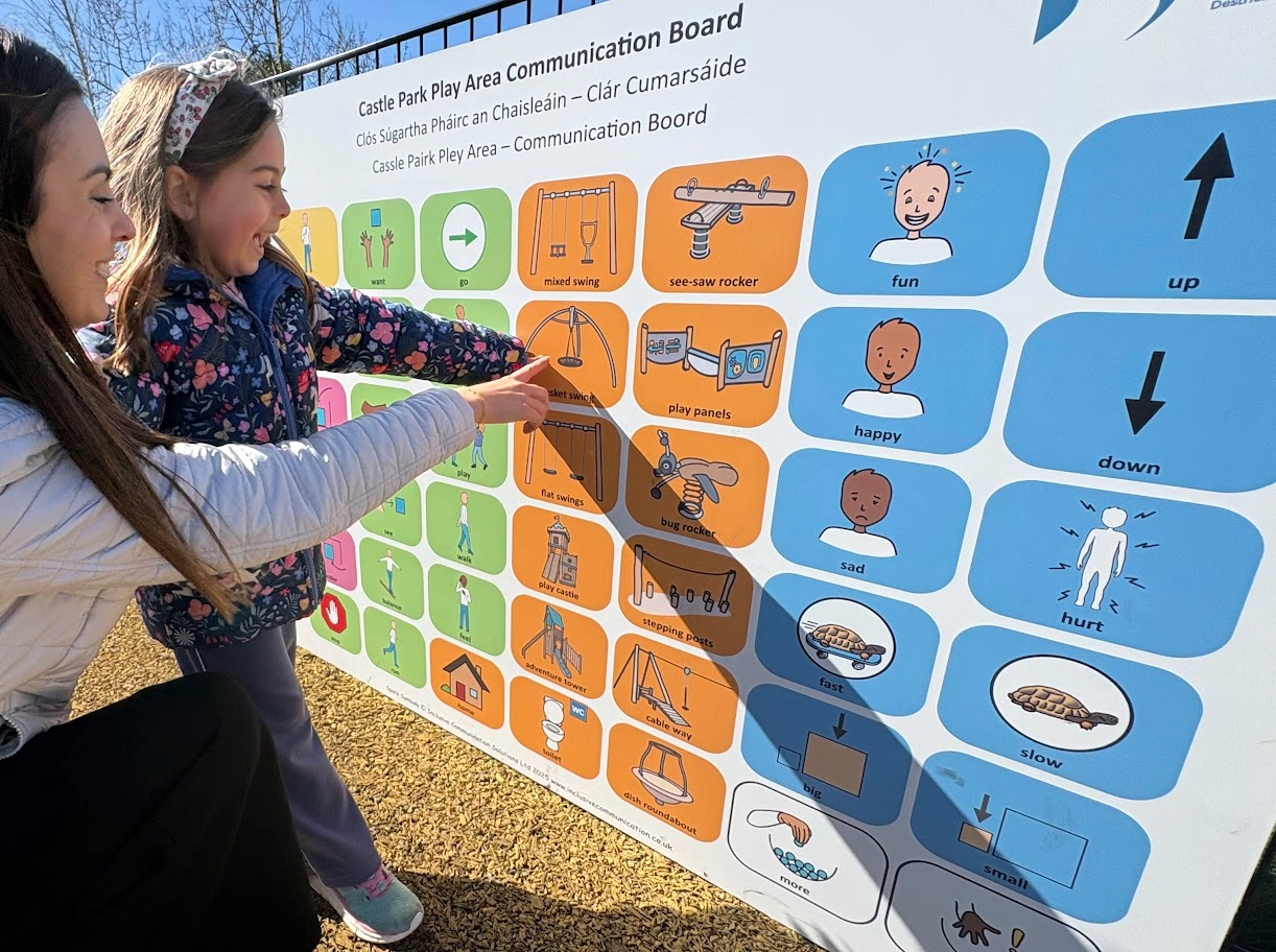 The photograph shows a young girl smiling and pointing to 'basket swing' on a large communication board. The girls Mum is standing next to her and is also pointing to the same symbol. The communication board is in Castle Park play area in Castlederg.