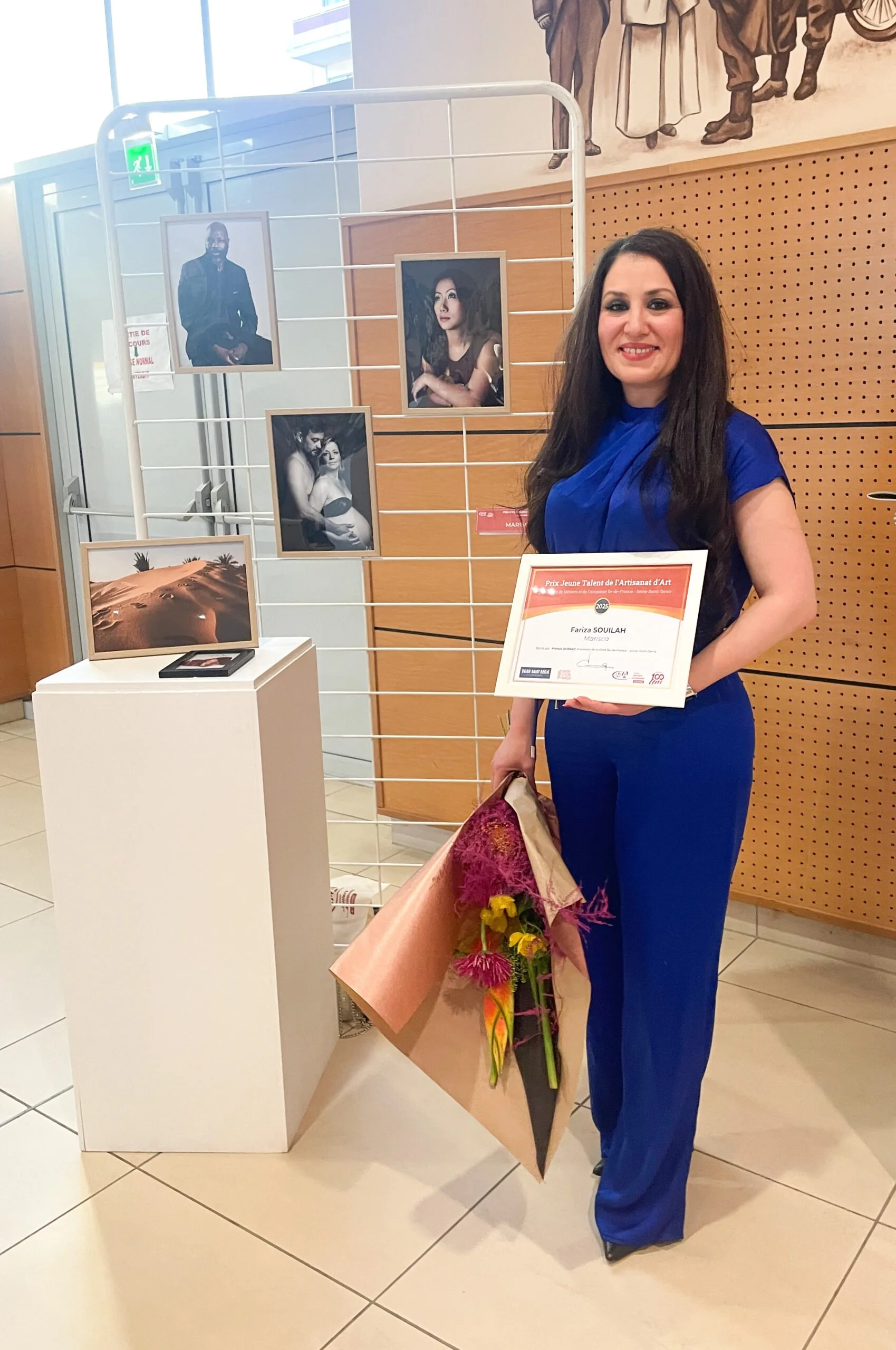 Une femme souriante portant une robe bleue, tenant une bouquet de fleurs et un certificat de reconnaissance, se tenant à côté d'une présentation photographique lors d'une cérémonie ou d'une exposition.
