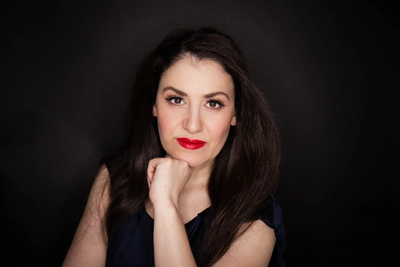 Portrait d'une femme avec des cheveux bruns, portant du rouge à lèvres rouge, posant contre un fond noir.