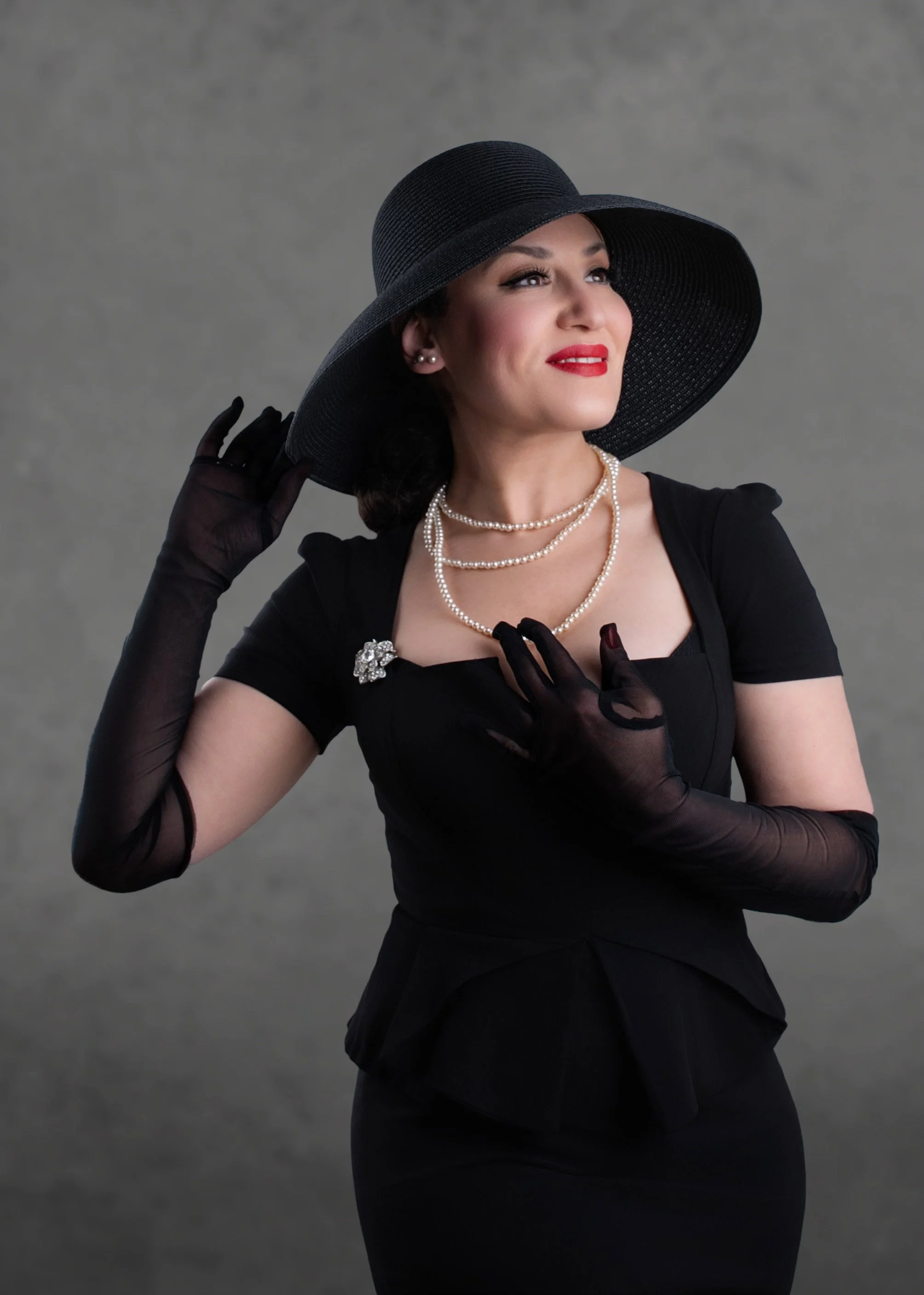 Femme élégante portant un chapeau noir, des gants en dentelle noire, un bijou en broche argentée, des colliers de perles, vêtue en noir, posant devant un fond gris.