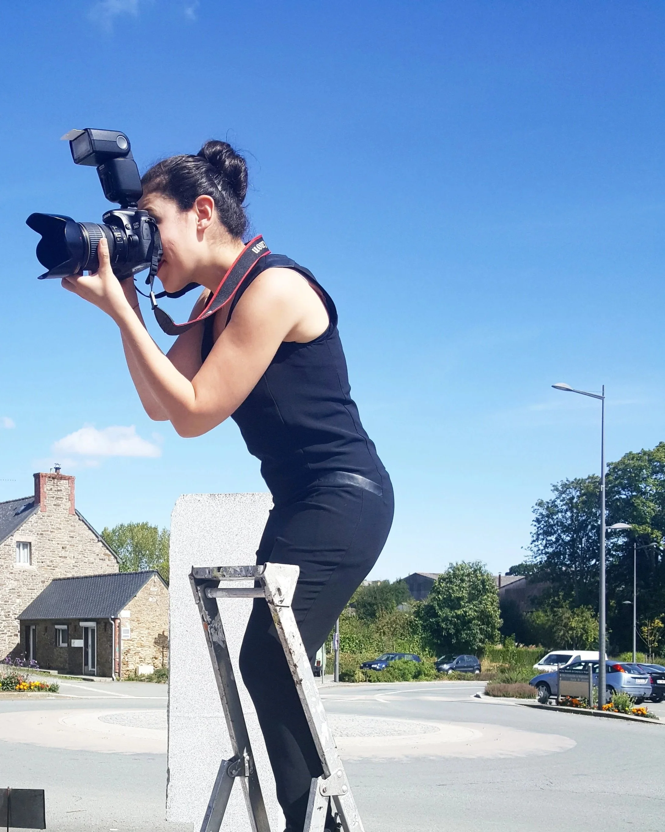 Une femme en tenu noir, se tenant sur une échelle, prenant une photo avec un appareil photo professionnel contre un ciel bleu clair.