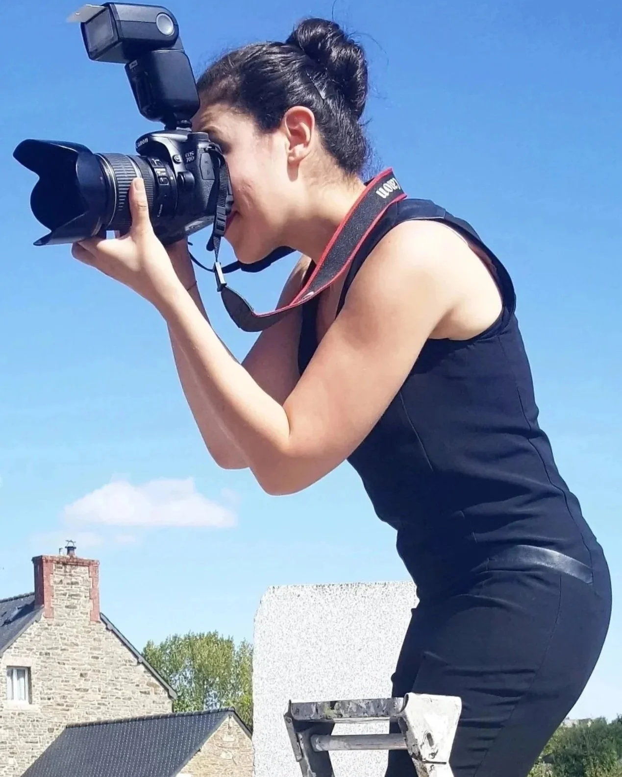 Une femme prenant une photo avec un appareil photo numérique, debout sur une échelle, contre un ciel clair et quelques maisons en arrière-plan.