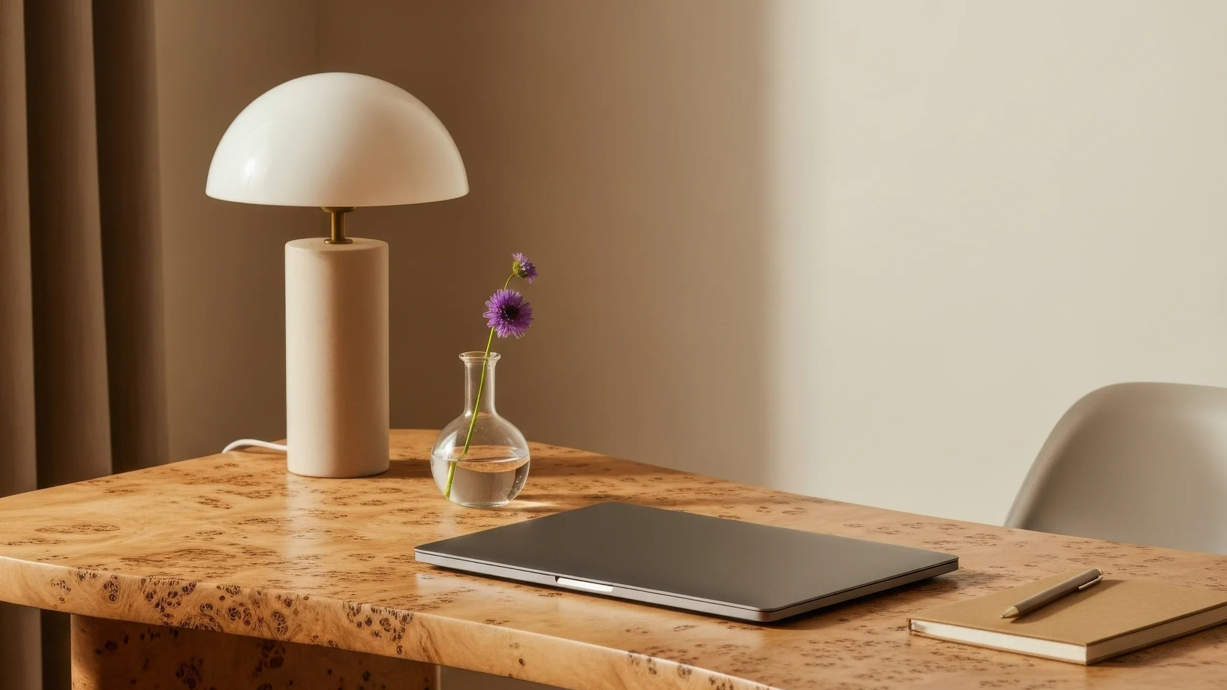 A wooden table with a modern table lamp, a small glass vase with purple flowers, a closed laptop, a closed notebook, and a pen.