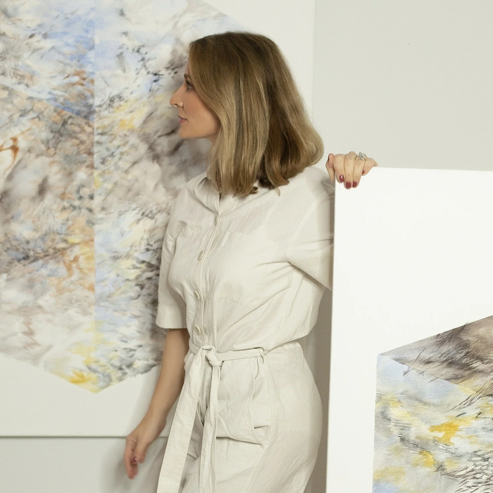 A woman with shoulder-length brown hair, wearing a light-colored dress, is standing indoors next to some abstract artwork.