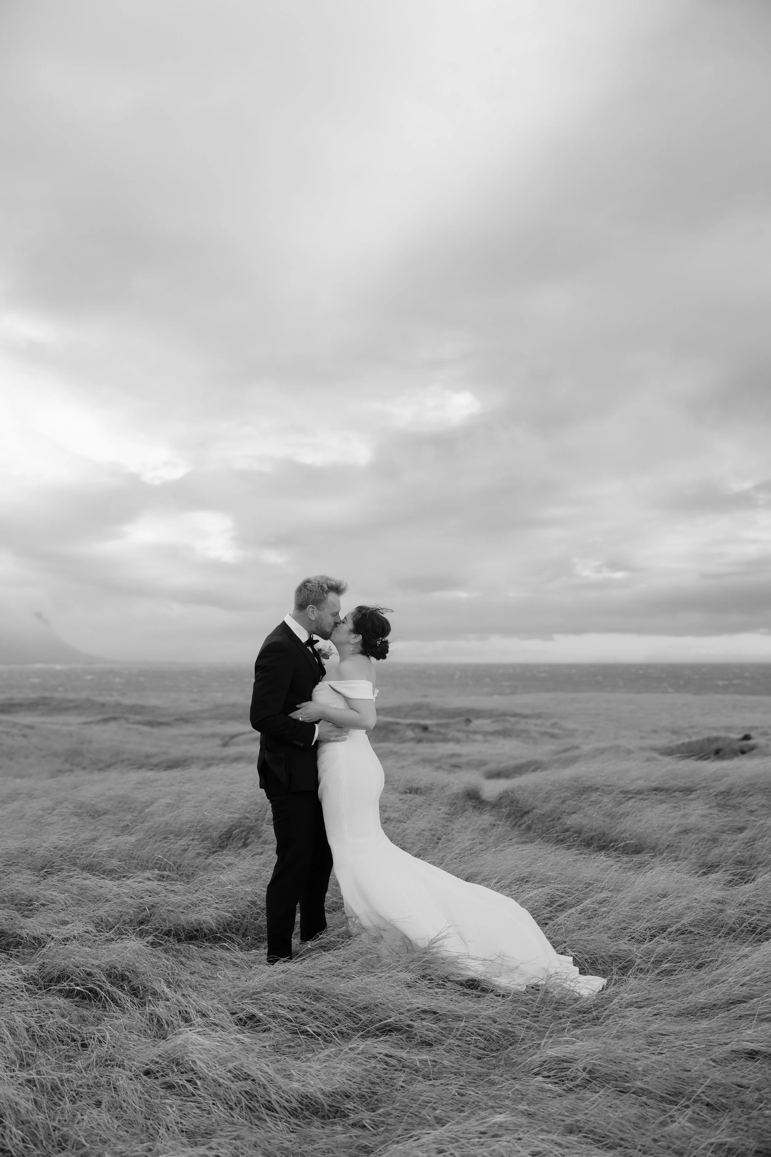 A black and white photo of a bride and groom in wedding attire sharing a kiss outdoors amid tall grass with a cloudy sky in the background during Iceland elopement.