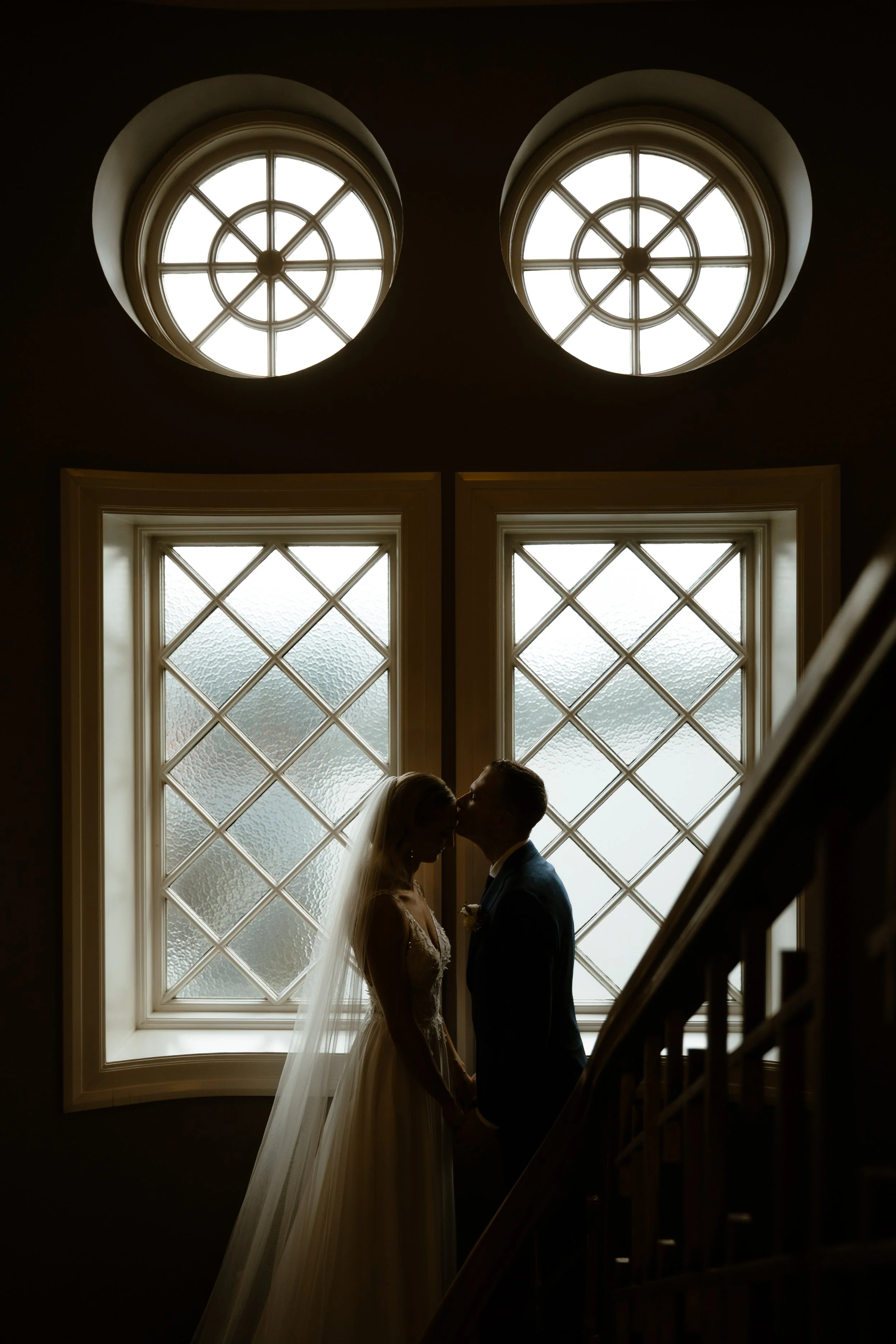 Silhouetted bride and groom standing close, holding hands, and about to kiss in front of large stained glass windows during an Iceland wedding photoshoot.