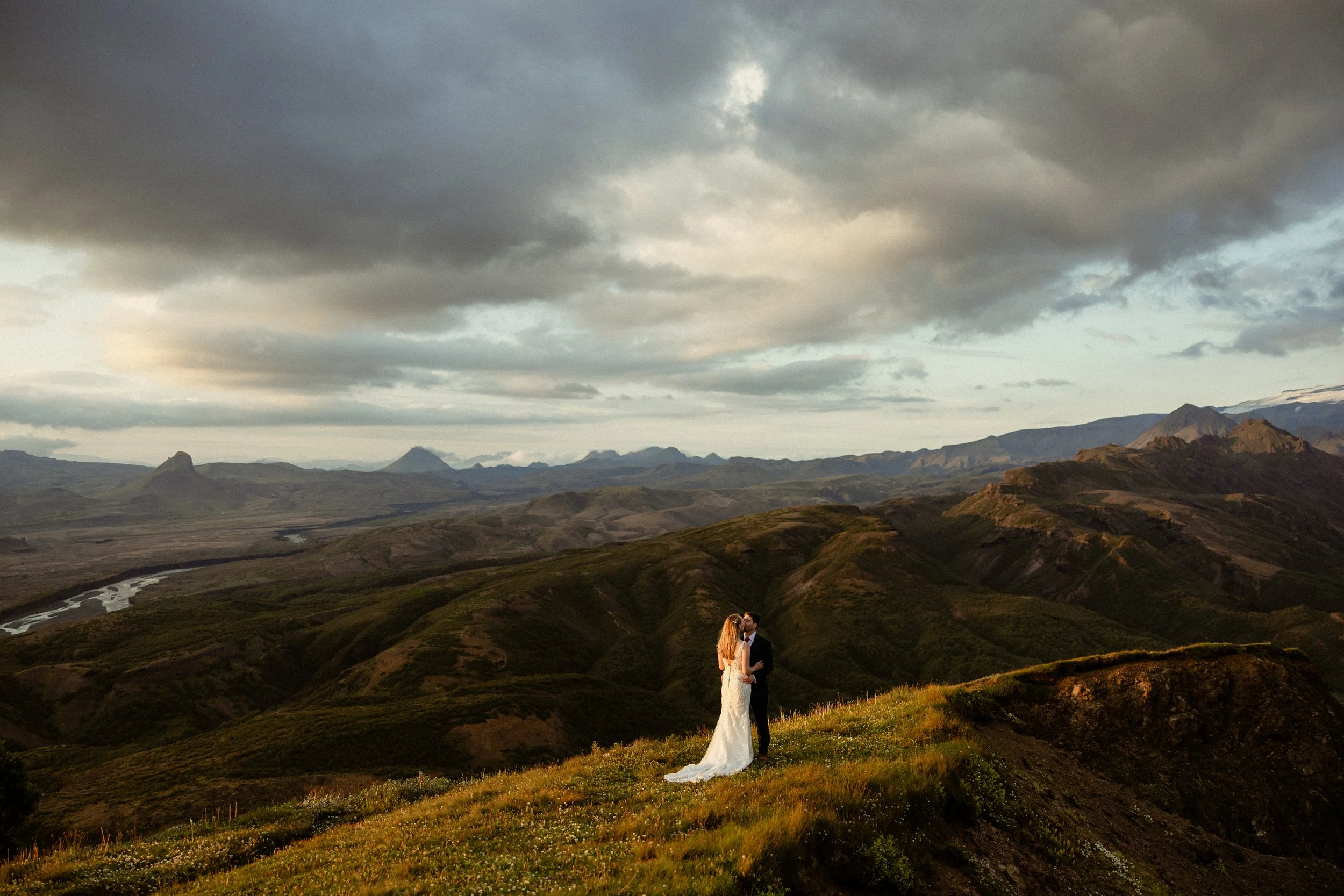 A couple standing on a grassy hill with mountains in the background, under a cloudy sky, with a bride in a white wedding dress and a groom in a dark suit during iceland wedding photos