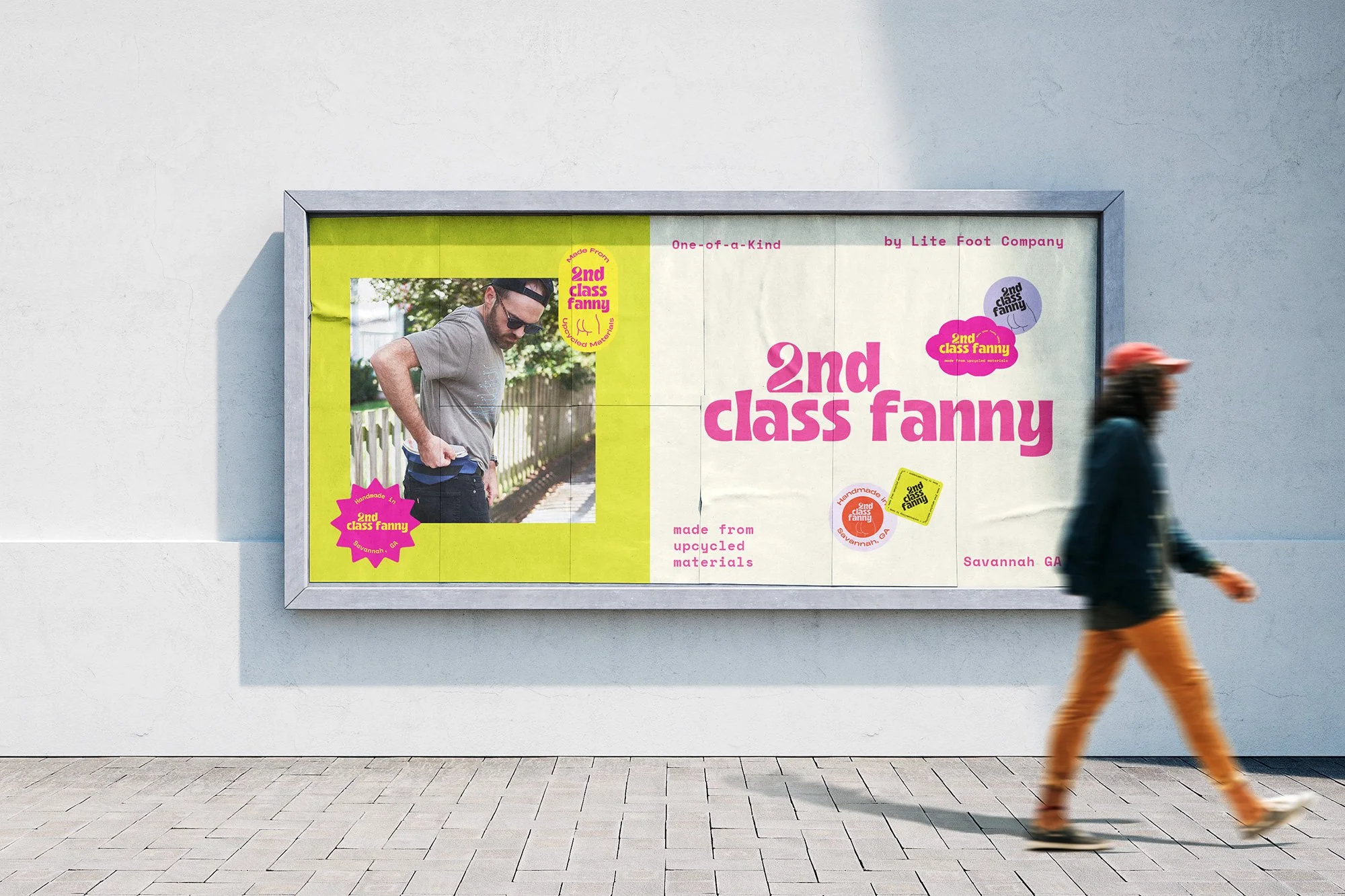 An outdoor billboard advertising 2nd class fanny, a product made from upcycled materials, featuring a man in a gray t-shirt and sunglasses holding a compact object and looking downward. The billboard has bright pink and yellow design elements with text and logos, located on a plain white wall.