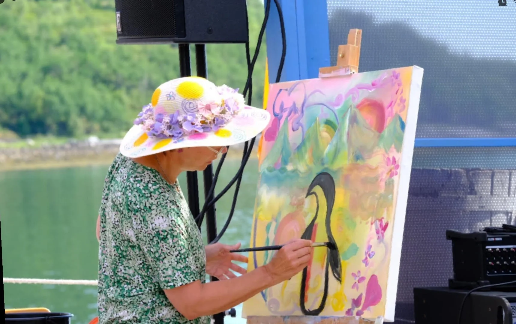 En kvinne i grønnblomstret bluse og stor stråhatt med fargerike blomster på står og maler en pingvin på et fargerikt maleri. Bakgrunnen er natur, skog og hav. Bildet av pingvinen er fargerikt.