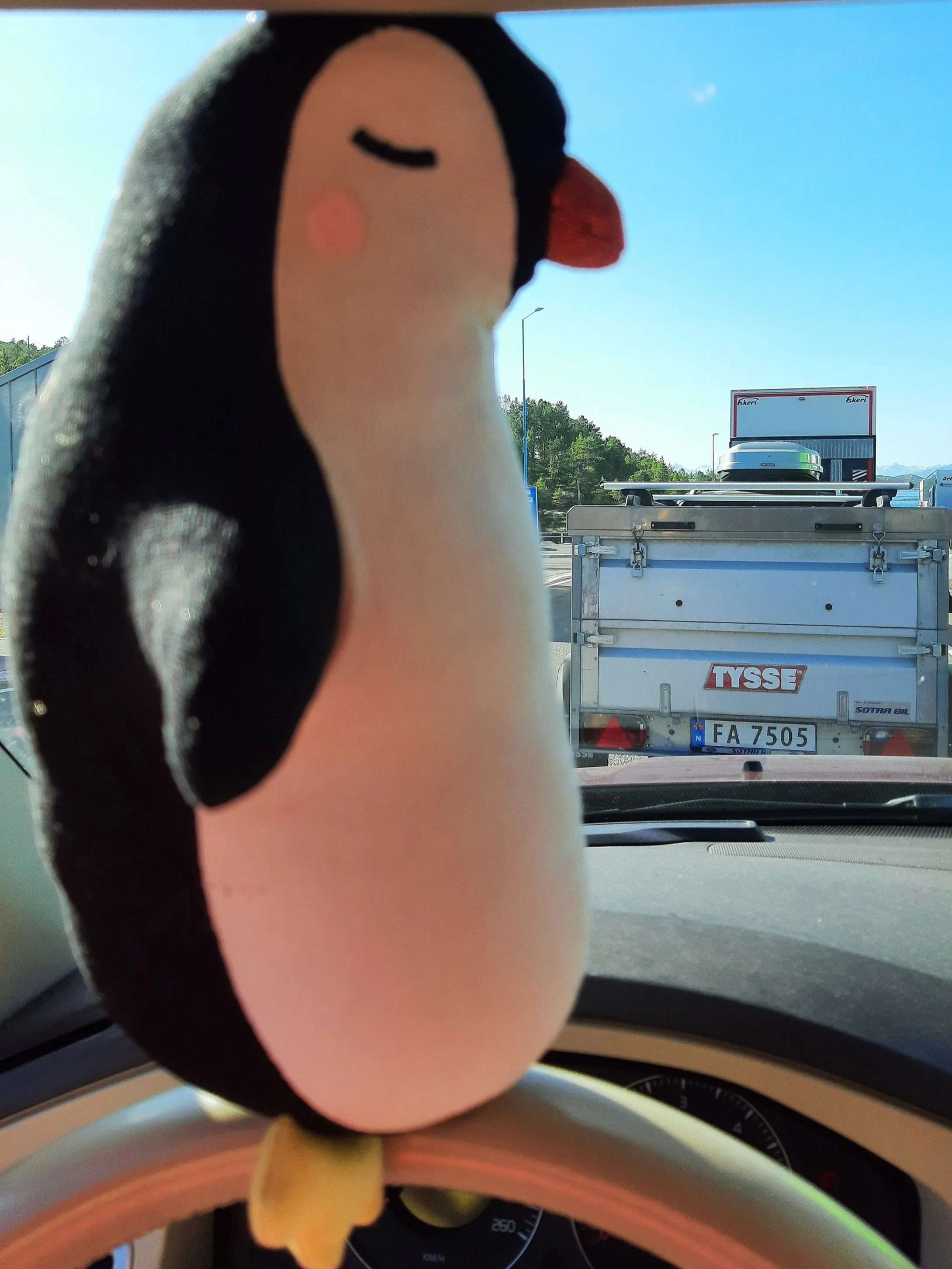 Vi ser en tøypingvin som sitter i en bilkø. Det er blå himmel i bakgrunnen og noen bilder. Pingvinen har lukkede øyne og ser fornøyd ut