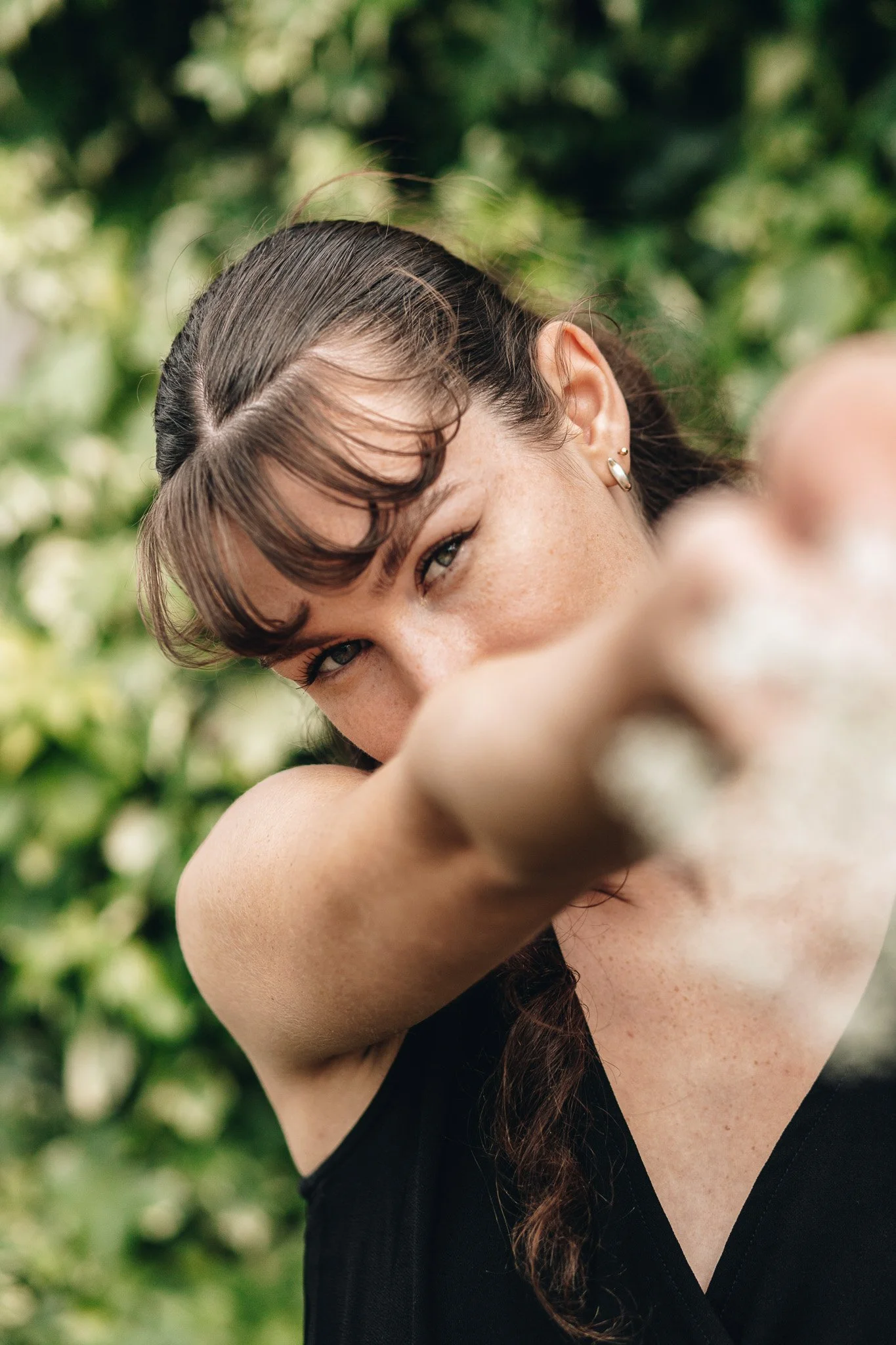 Close-up of a woman with brown hair, wearing earrings and a black sleeveless top, with her arm across her face, outdoors with green foliage in the background.